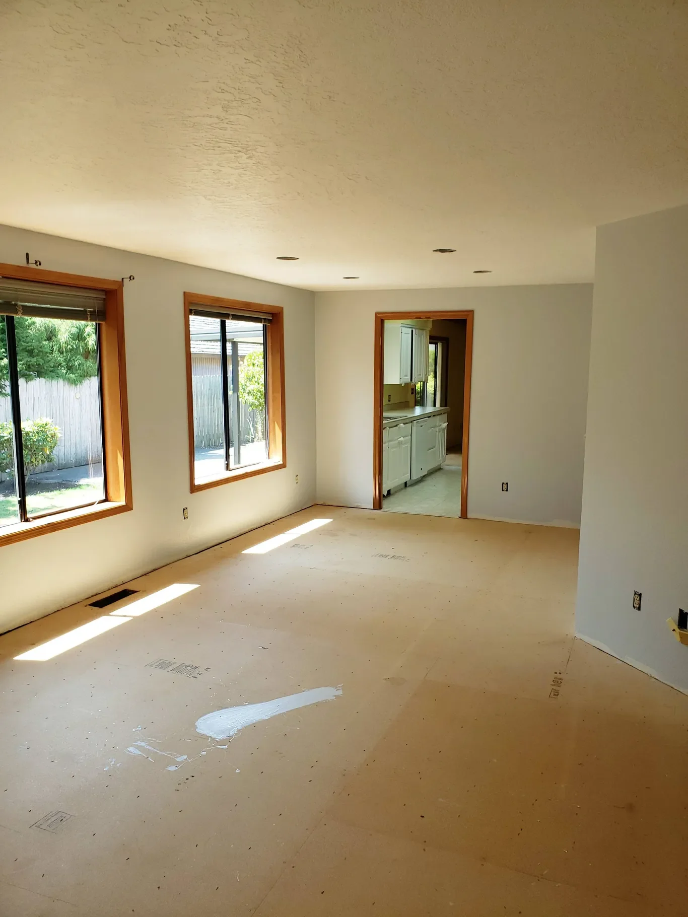 Empty living room with large windows, light-colored walls, and an open doorway leading to a kitchen with white cabinets.