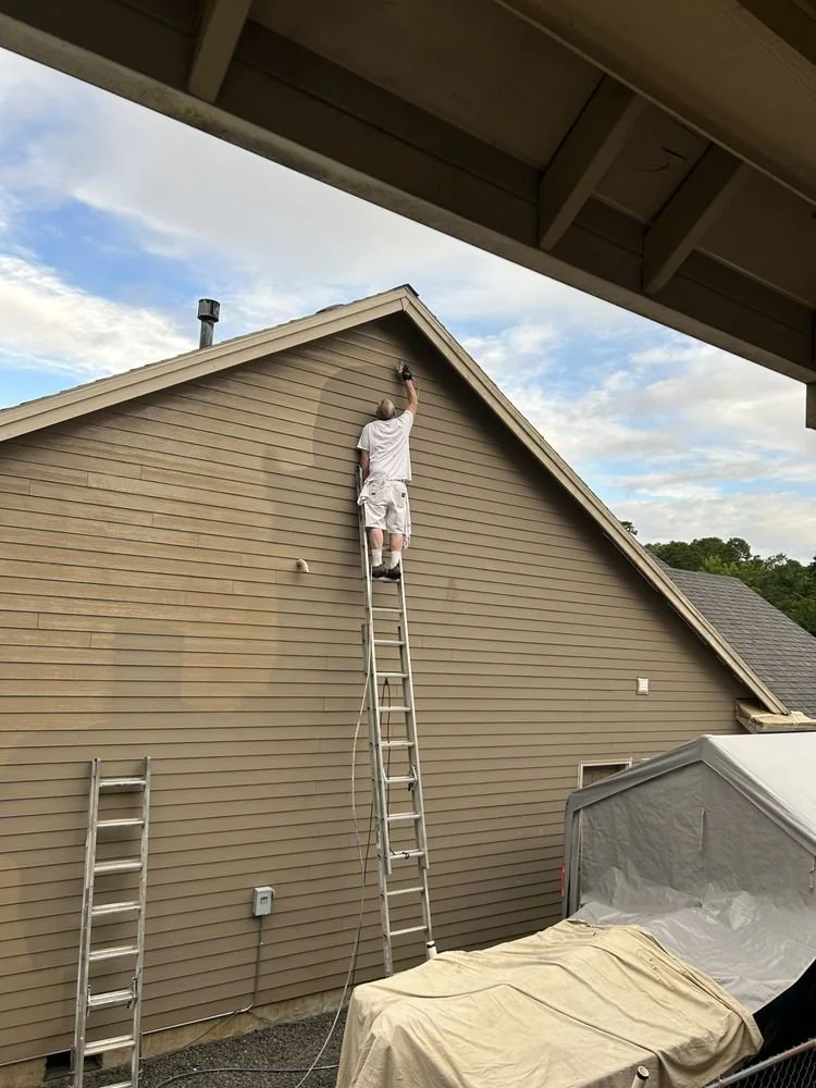 A man is standing on a ladder painting the upper part of a house's exterior wall under the roof. There is another ladder leaning against the wall and a vehicle covered by a tarp nearby. The sky is partly cloudy.