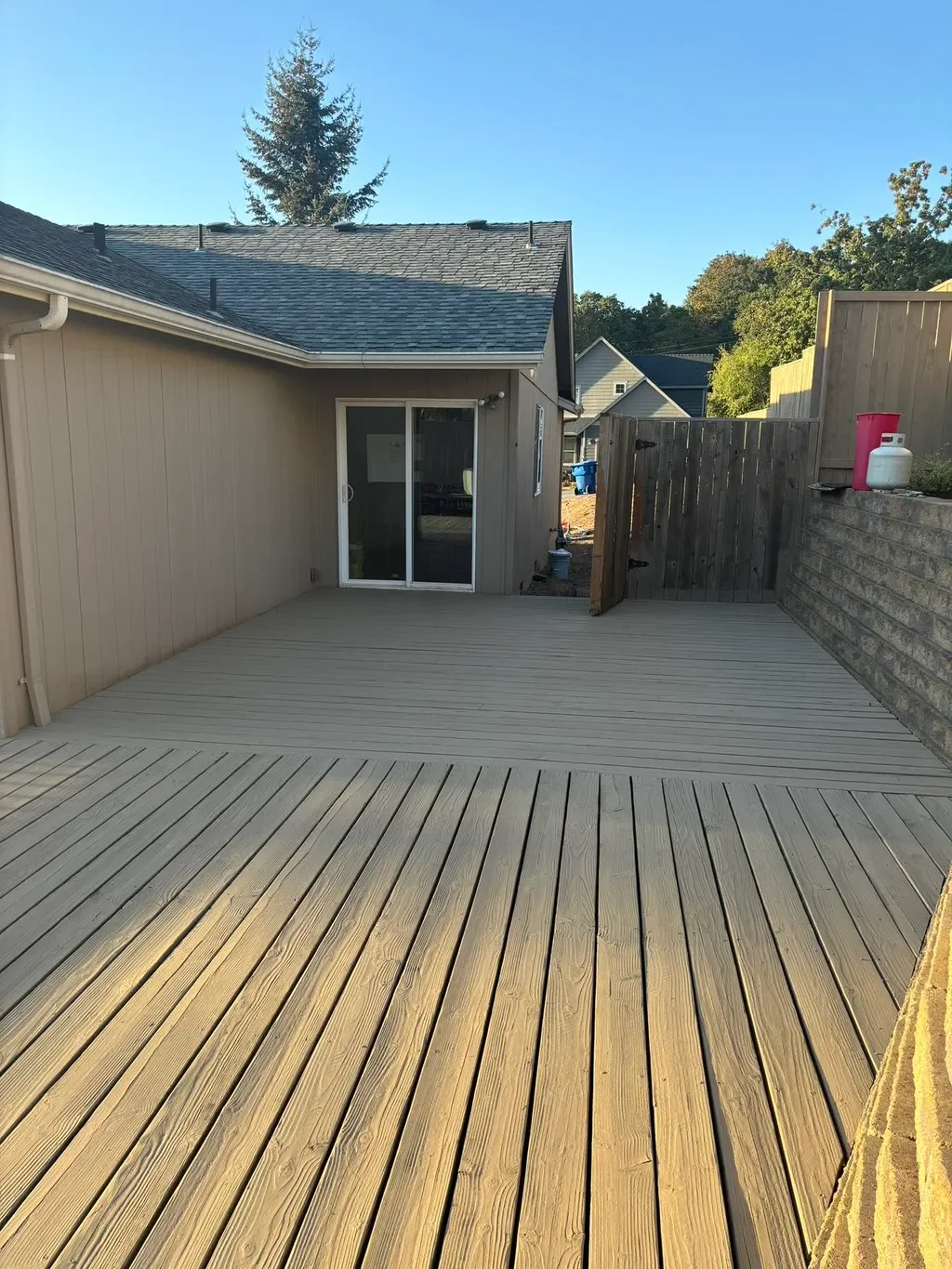 A newly constructed wooden deck outside a house, with a sliding glass door and a partially open wooden gate, surrounded by neighboring houses and greenery.