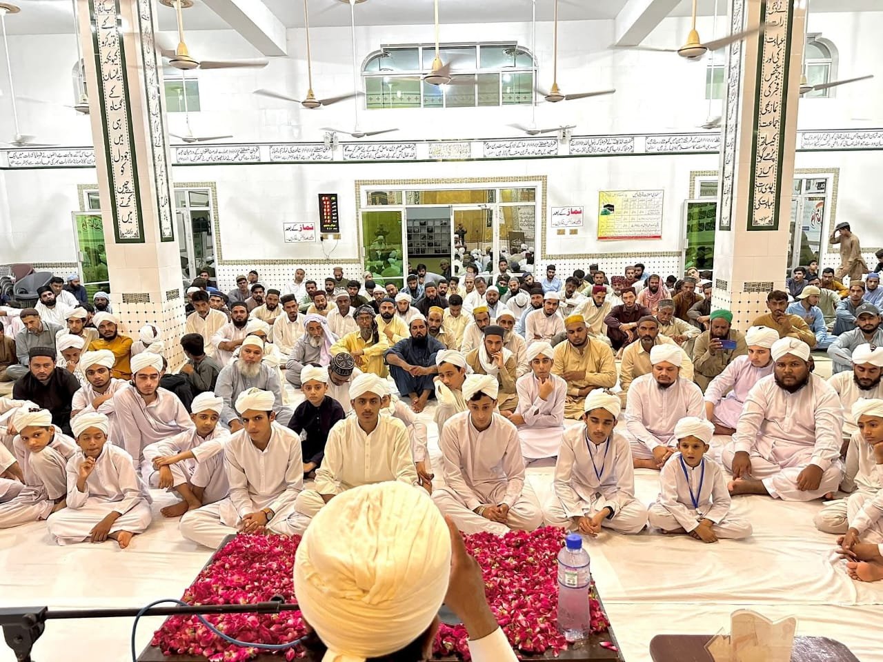 Group of Muslim men and boys sitting on the floor in a mosque, facing the speaker, who is seated with their back to the camera, with pink flowers and a water bottle in front. The mosque has white walls with Arabic calligraphy and ceiling fans.
