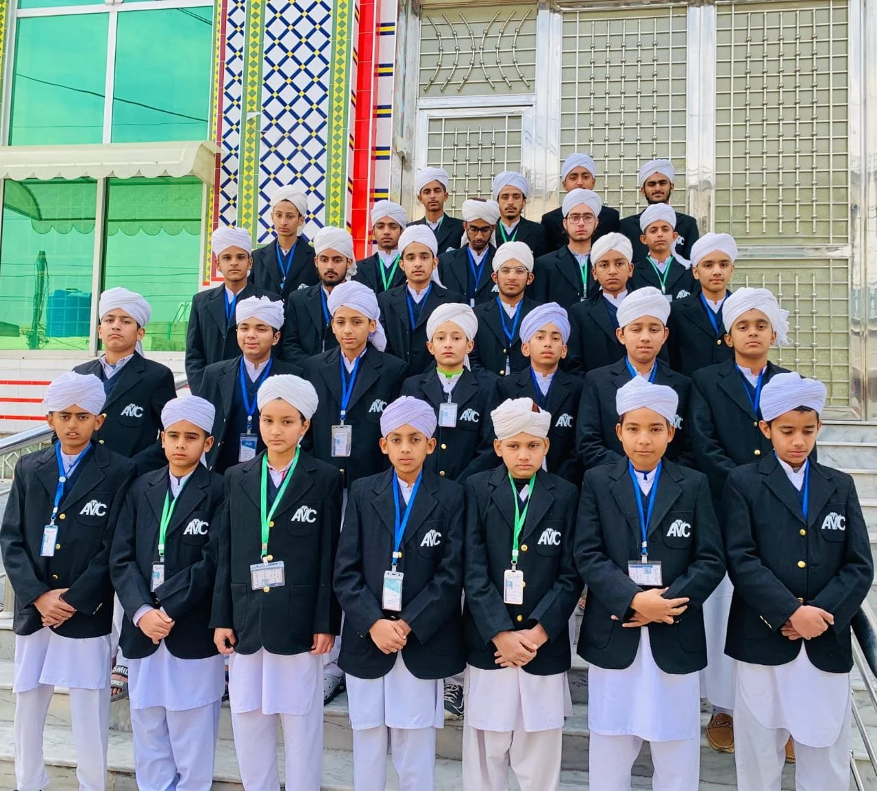 Group of young boys and teenagers wearing black blazers, white turbans, and white pants, standing on stairs in front of a building with colorful decorative tiles and green windows, posing for a group photo.