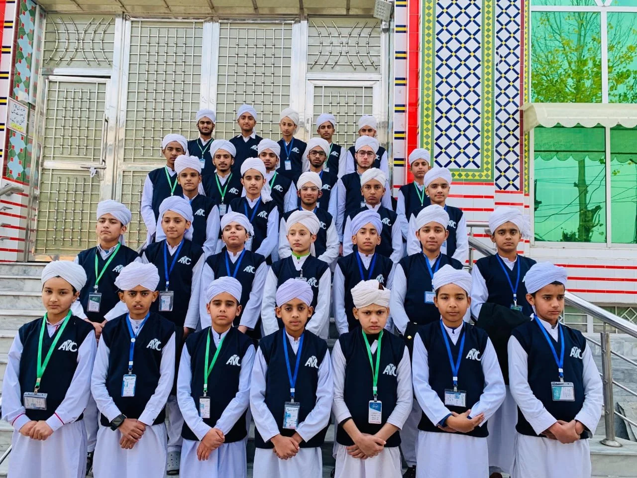 Group of young students dressed in traditional white turbans and matching navy blue vests with white shirts, standing on steps outside a building with colorful geometric and floral designs.