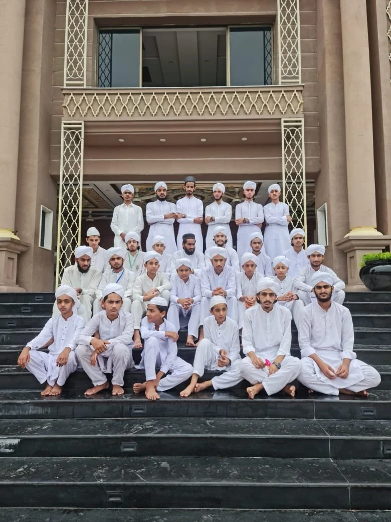 Group of young men dressed in white traditional Islamic attire and caps, sitting and standing on stairs outside a building with ornate railings and columns.