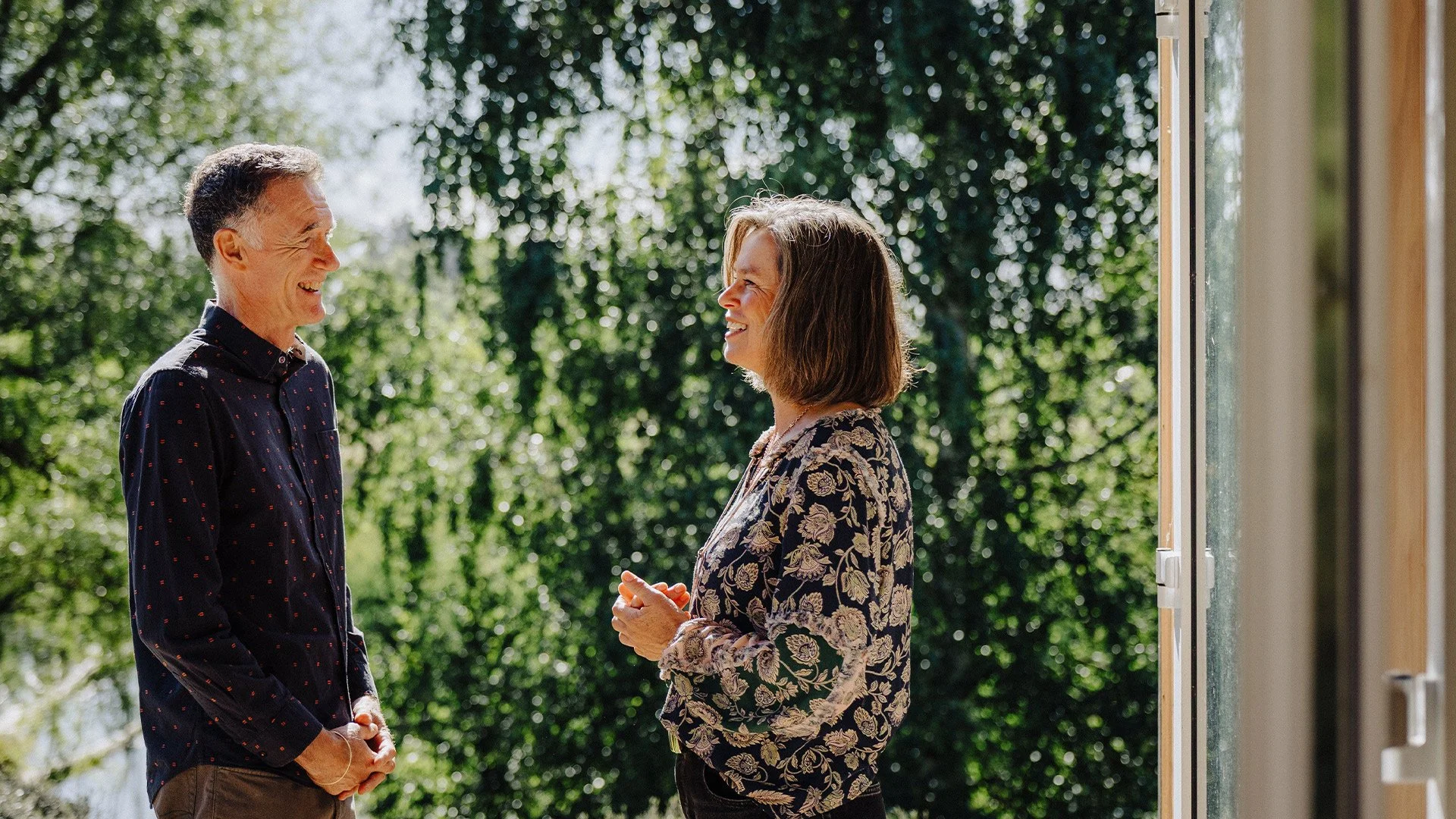 A man and woman smiling and talking outdoors on a sunny day, standing near an open door with trees in the background.