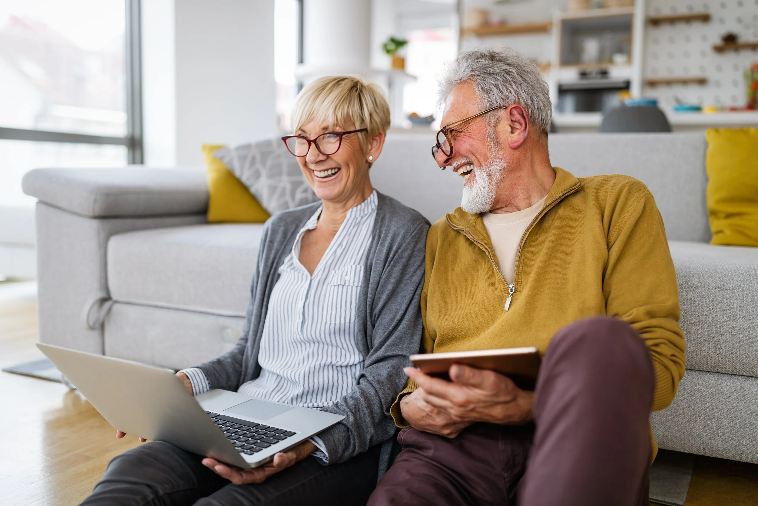 An elderly couple sitting on the floor in a bright living room, laughing while using a laptop and a tablet.
