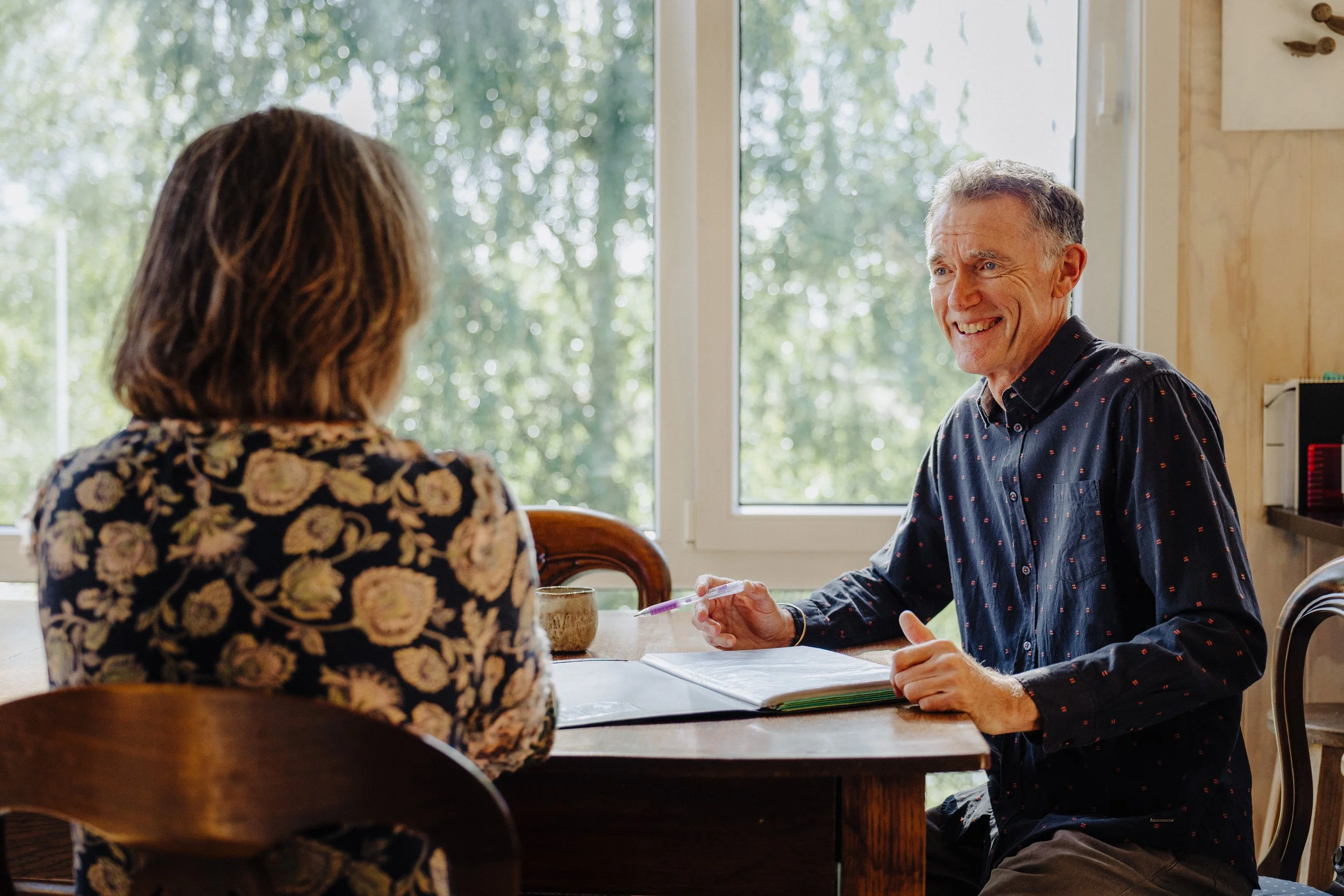 A man and woman sitting across from each other at a dining table, having a conversation indoors near a large window with trees outside.