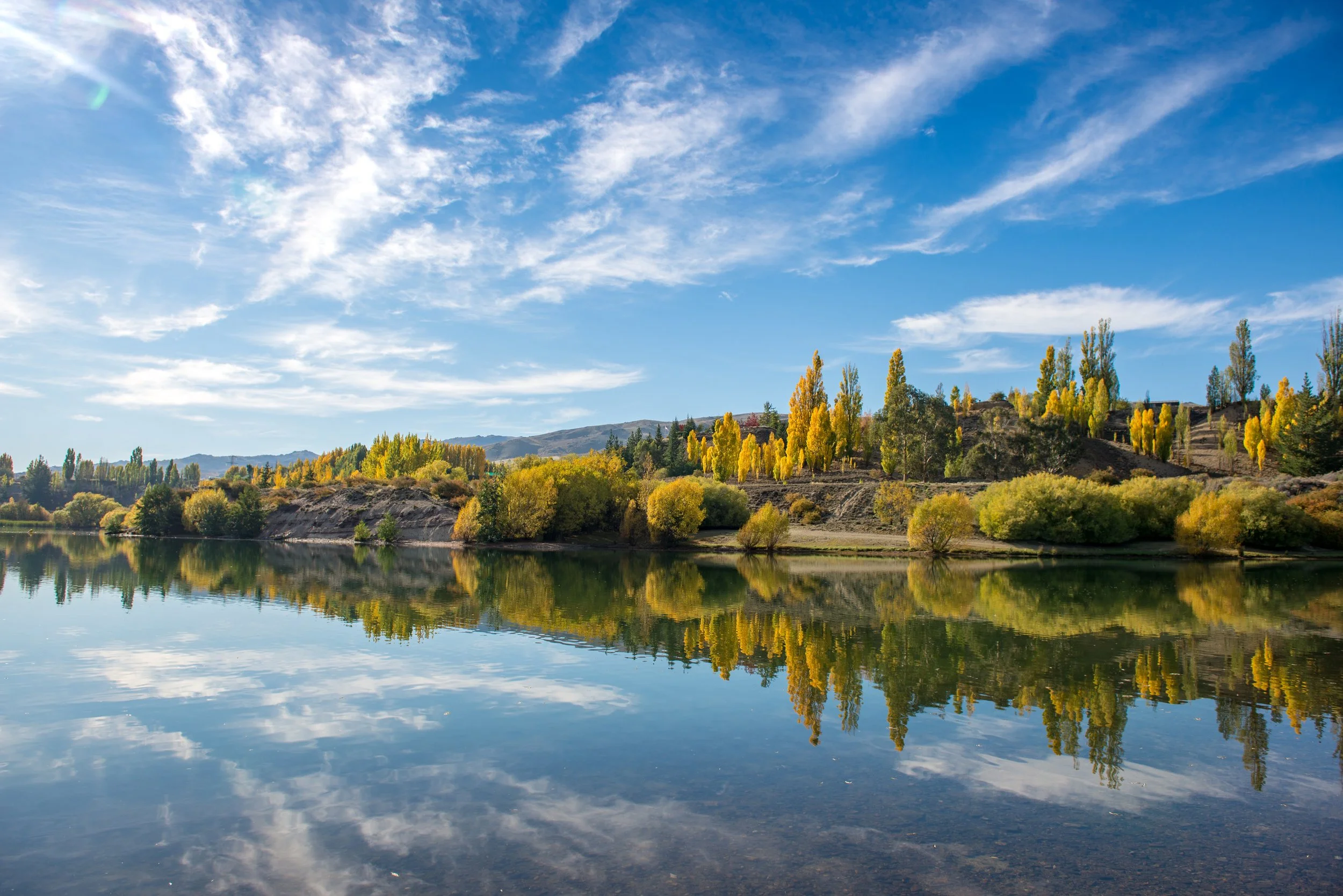 Scenic view of a lake with trees displaying fall foliage and mountains in the distance, under a blue sky with scattered clouds.