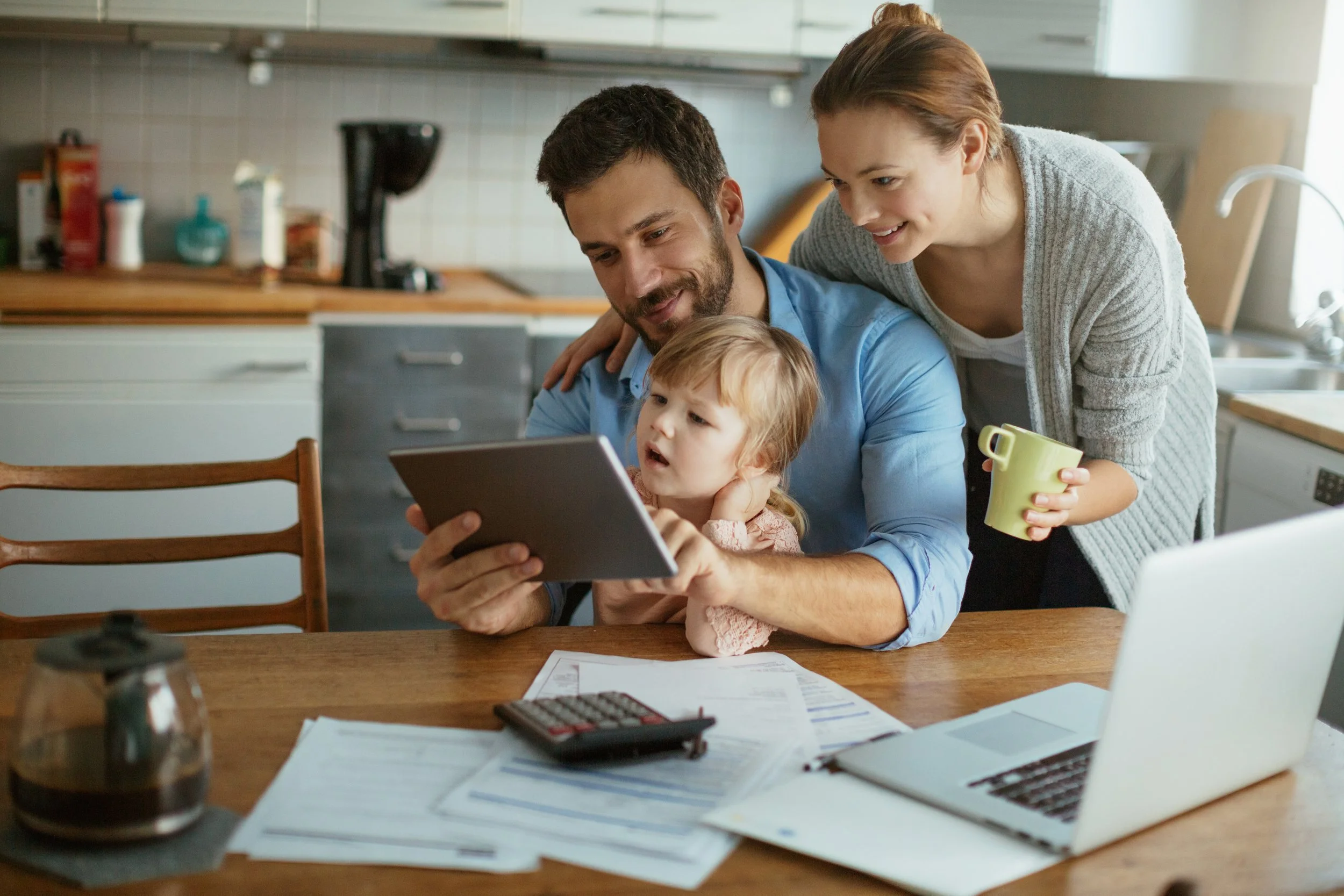 Family of three gathered around a table, looking at a tablet, with papers, a laptop, and a calculator in front of them, in a kitchen.