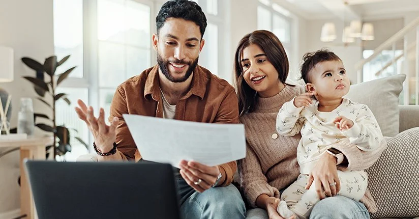 Family of three sitting on a couch in a bright living room, looking at a laptop and a document.