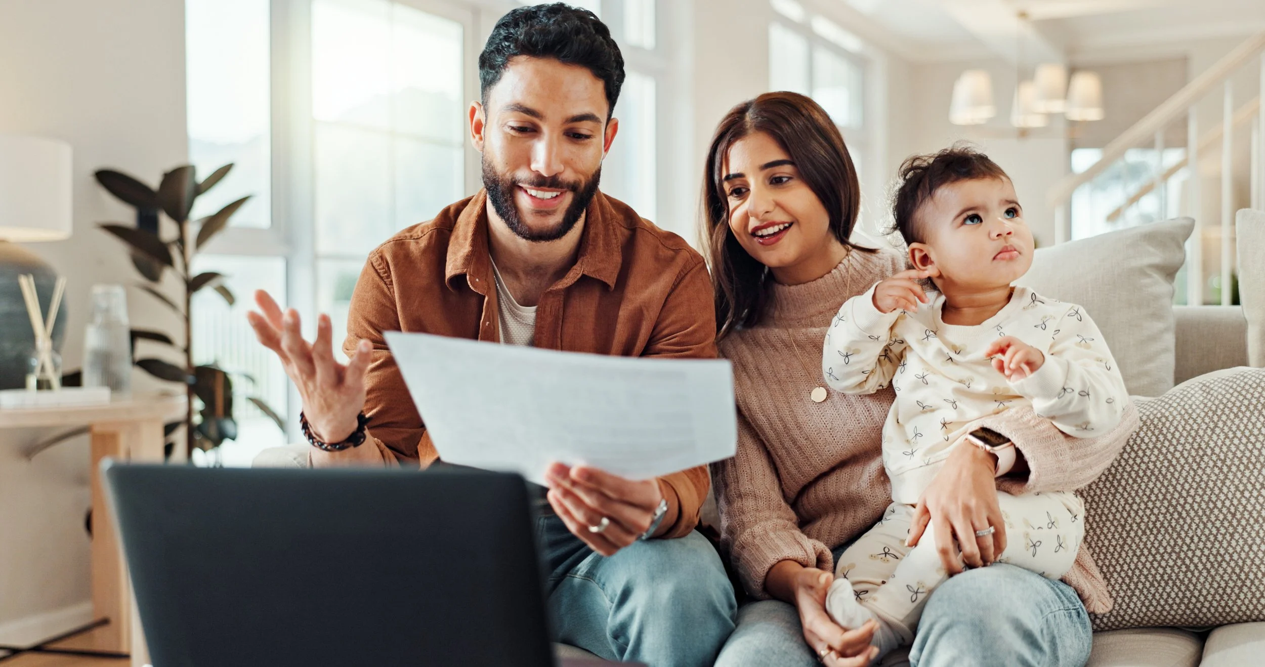 A family of three sitting on a beige couch in their living room, looking at a laptop and a document together. The man has dark hair and a beard, wearing a brown shirt. The woman has shoulder-length dark hair, wearing a beige sweater. Their toddler, with short dark hair, is sitting on the woman's lap, wearing pajamas with bow patterns and looking up thoughtfully.