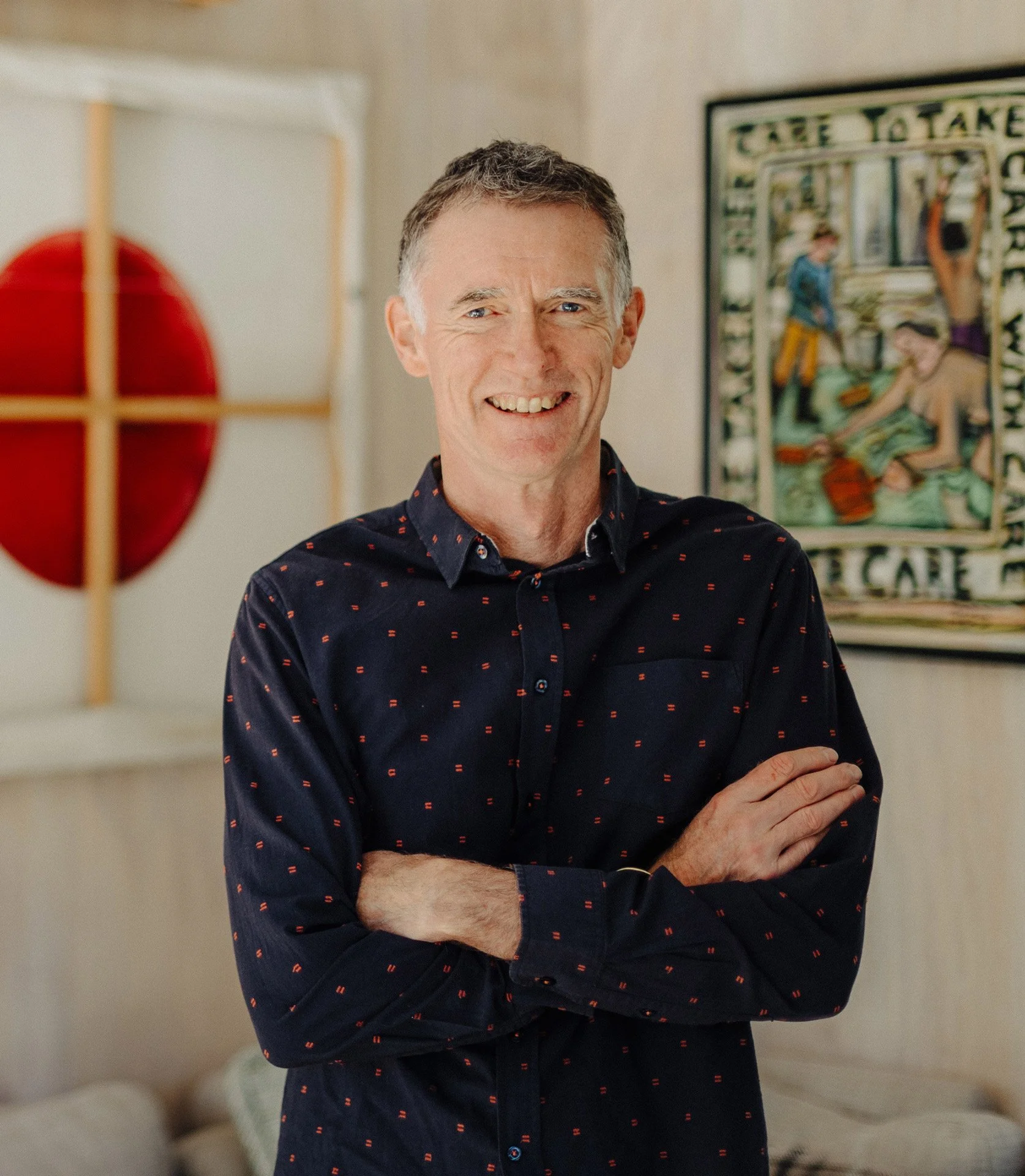 A middle-aged man with short, light brown hair standing indoors with his arms crossed, smiling at the camera. He is wearing a dark blue button-up shirt with small red patterns. In the background, there is a framed painting and a decorative wall hanging with a red circle and wooden cross design.