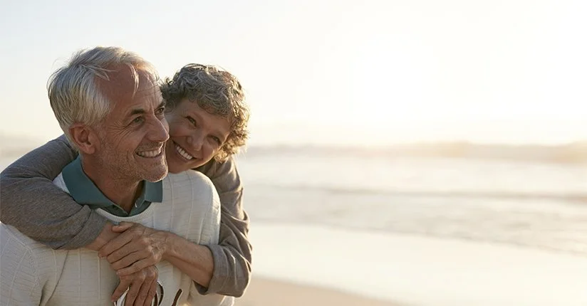 An older man and a woman smiling at the beach, with the woman hugging the man from behind.
