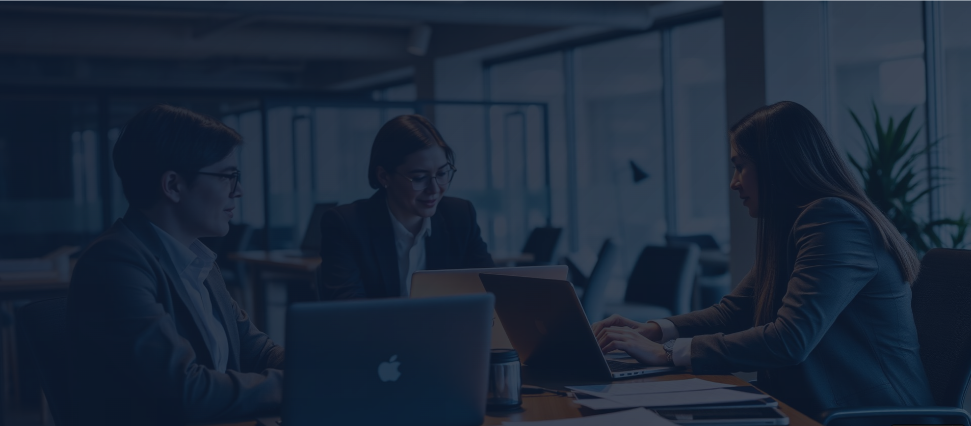 Three women working on laptops at a conference table in a modern office.