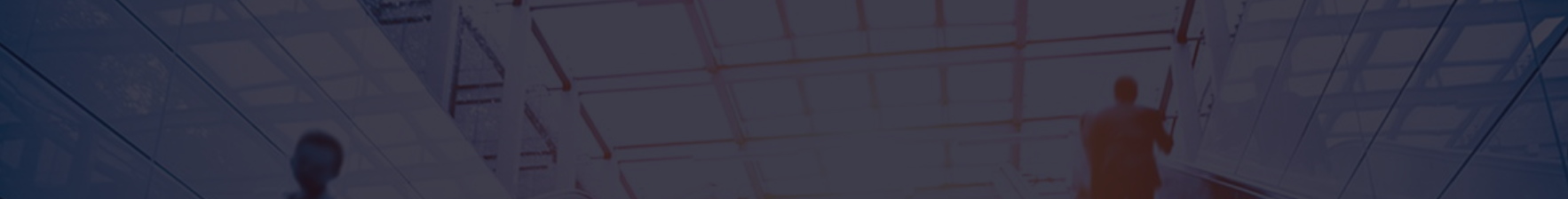 Industrial setting with a person working or inspecting equipment in a large, open warehouse or factory space, with overhead piping and structural elements visible.