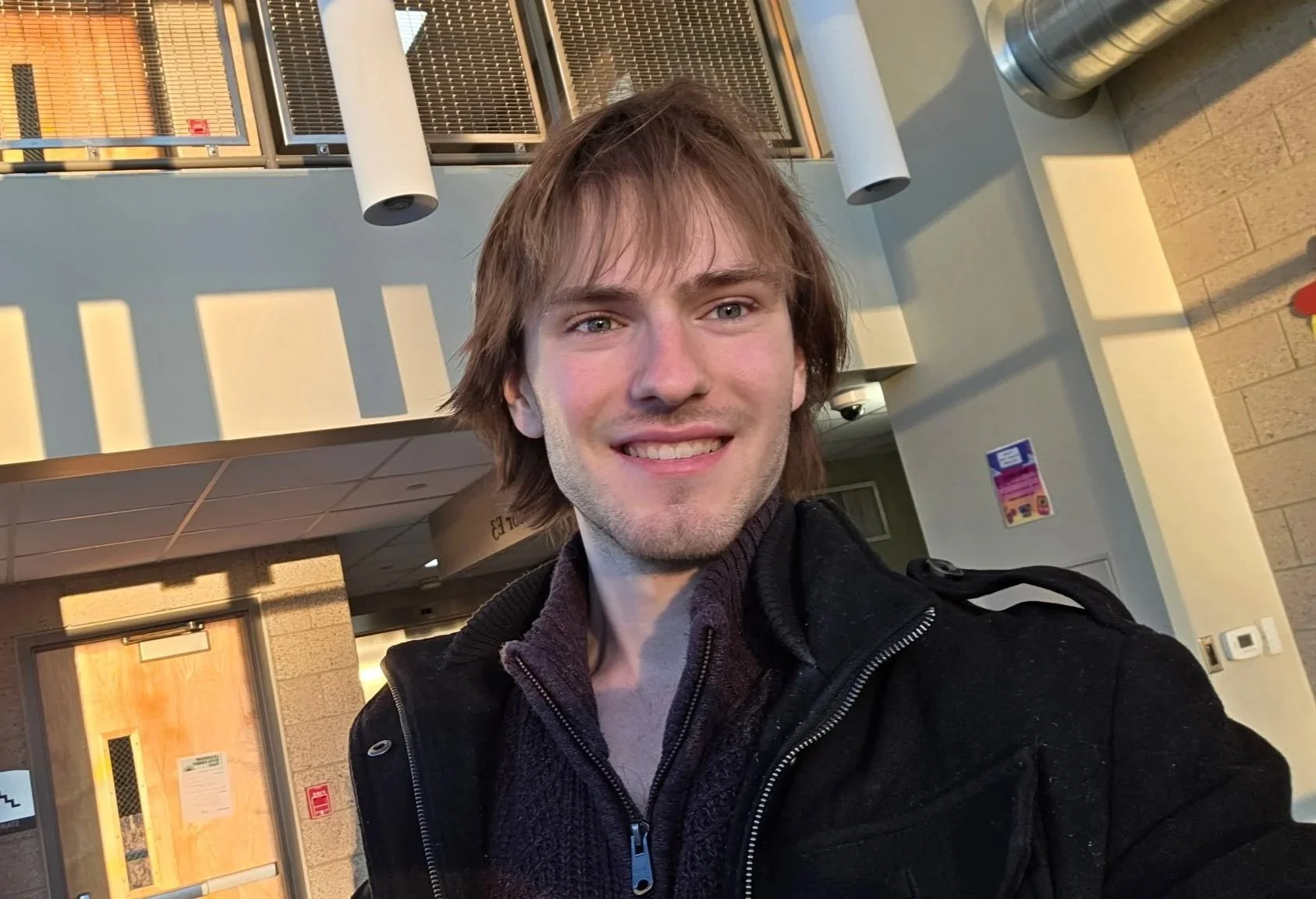 A young man with light brown hair and a big smile taking a selfie indoors, with sunlight casting shadows on his face, wearing a black jacket and a dark sweater, in a building with beige walls, ceiling lights, and ventilation ducts.
