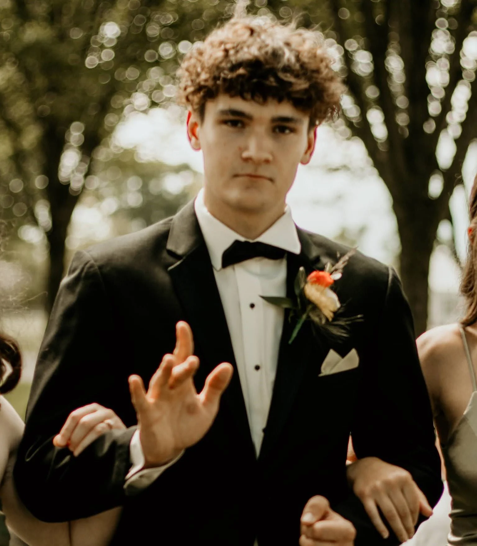 Young man in a black tuxedo with a corsage, raising one hand in a gesture, outdoors with trees in the background.