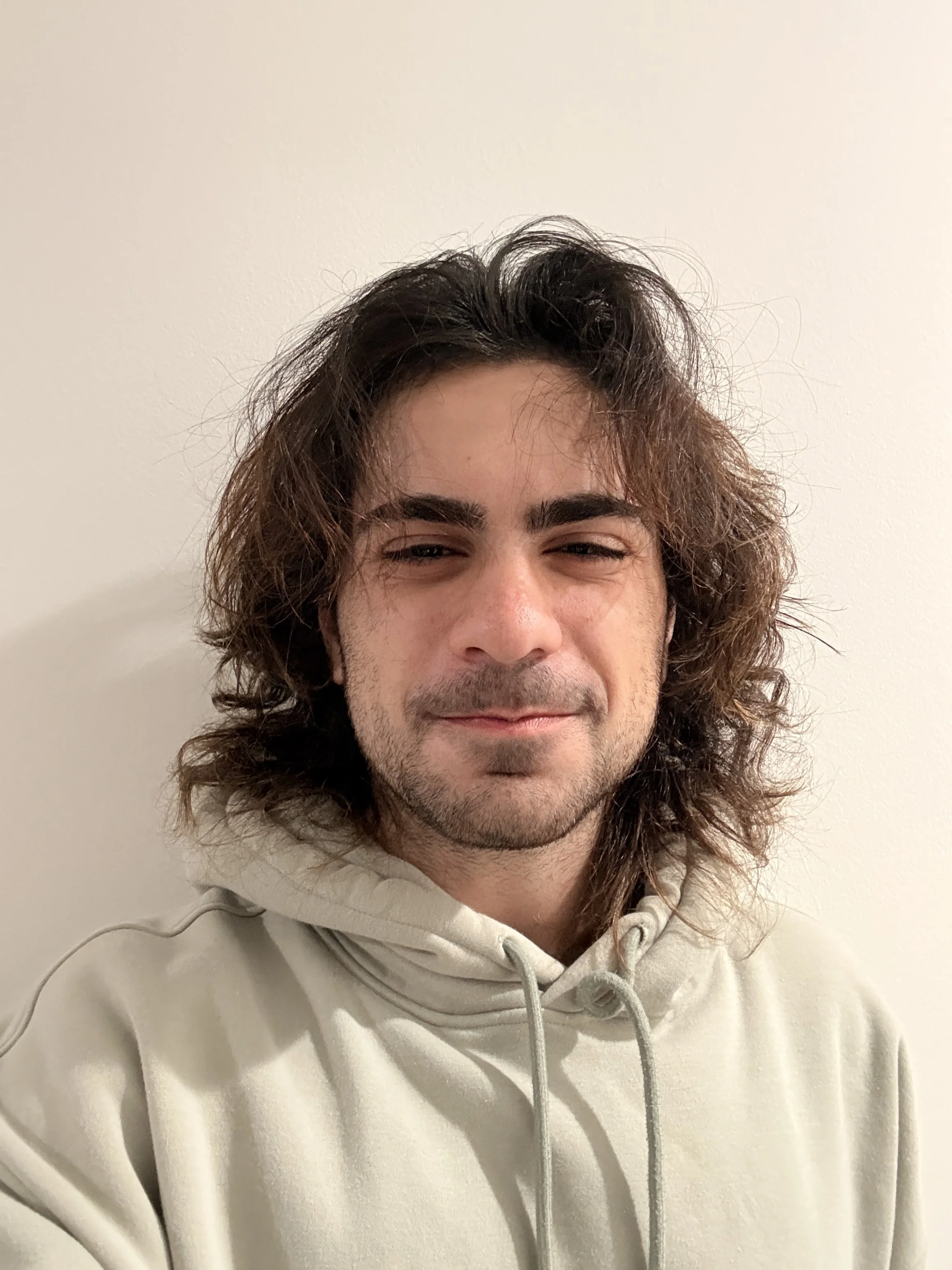 A young man with long, wavy brown hair and a light beard, wearing a light-colored hoodie, standing against a plain white wall.