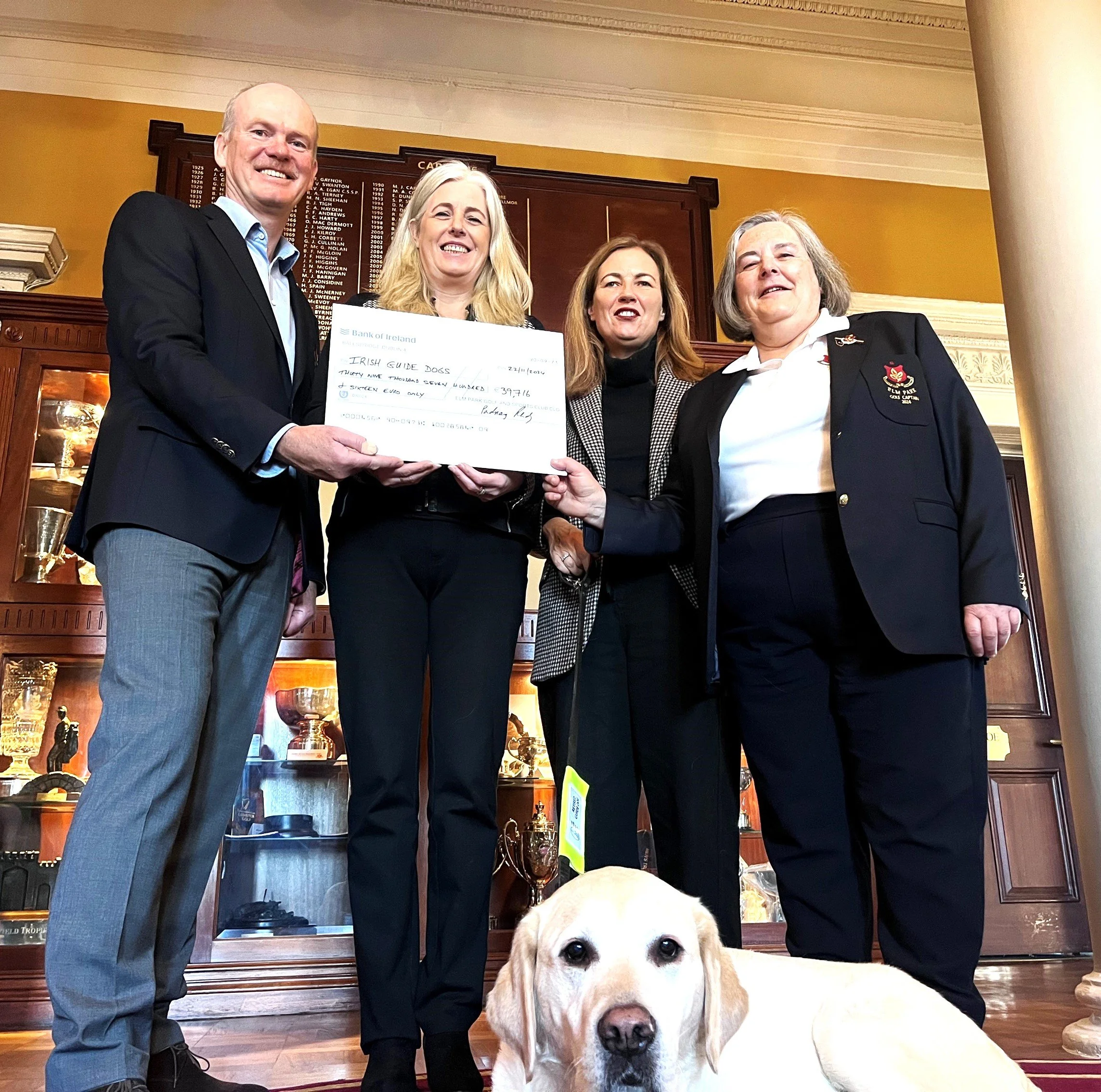 Four women and one man holding a large check together in a room with trophies behind them and a Labrador Retriever sitting in front.