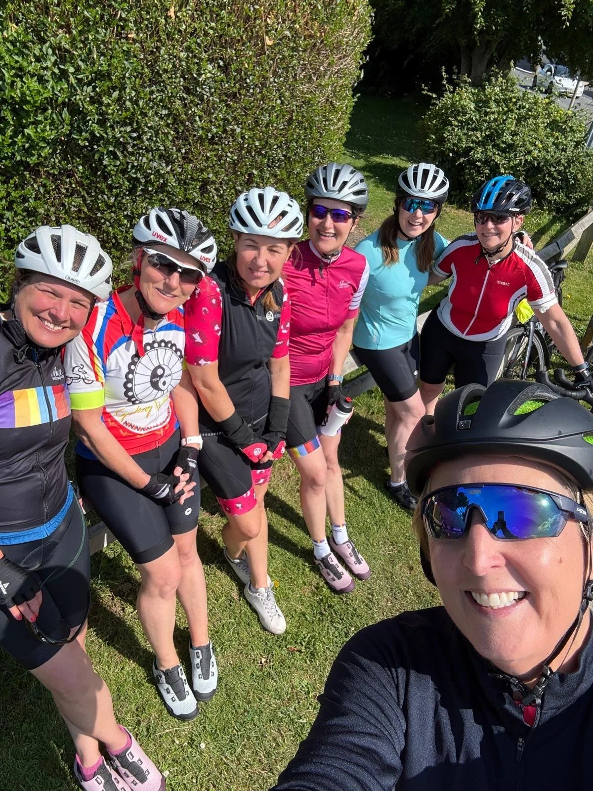 Group of six women in cycling gear and helmets taking a selfie on a grassy area with bushes and trees in the background.