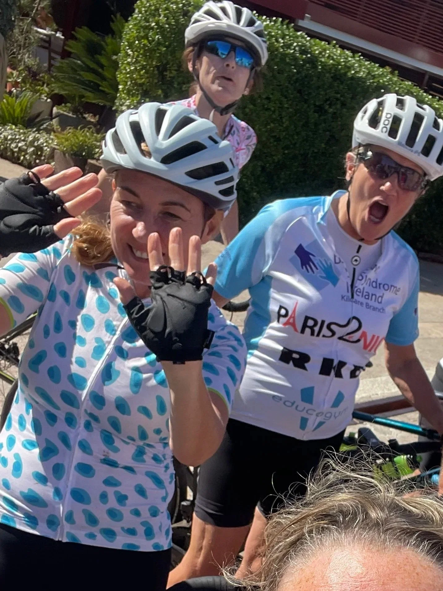 Three women in cycling gear and helmets posing for a photo, with two women making a playful gesture and one woman shouting or singing, outdoors with greenery in the background.