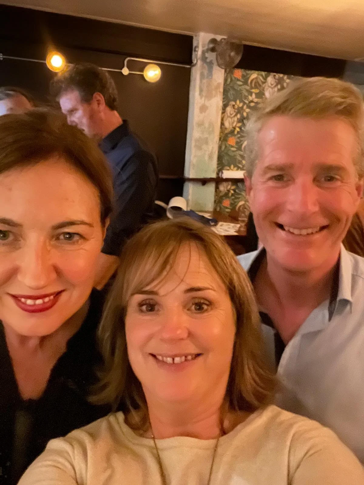 Three people taking a selfie at a dimly lit indoor gathering, smiling at the camera, with a woman in the center, and a woman on the left and a man on the right.