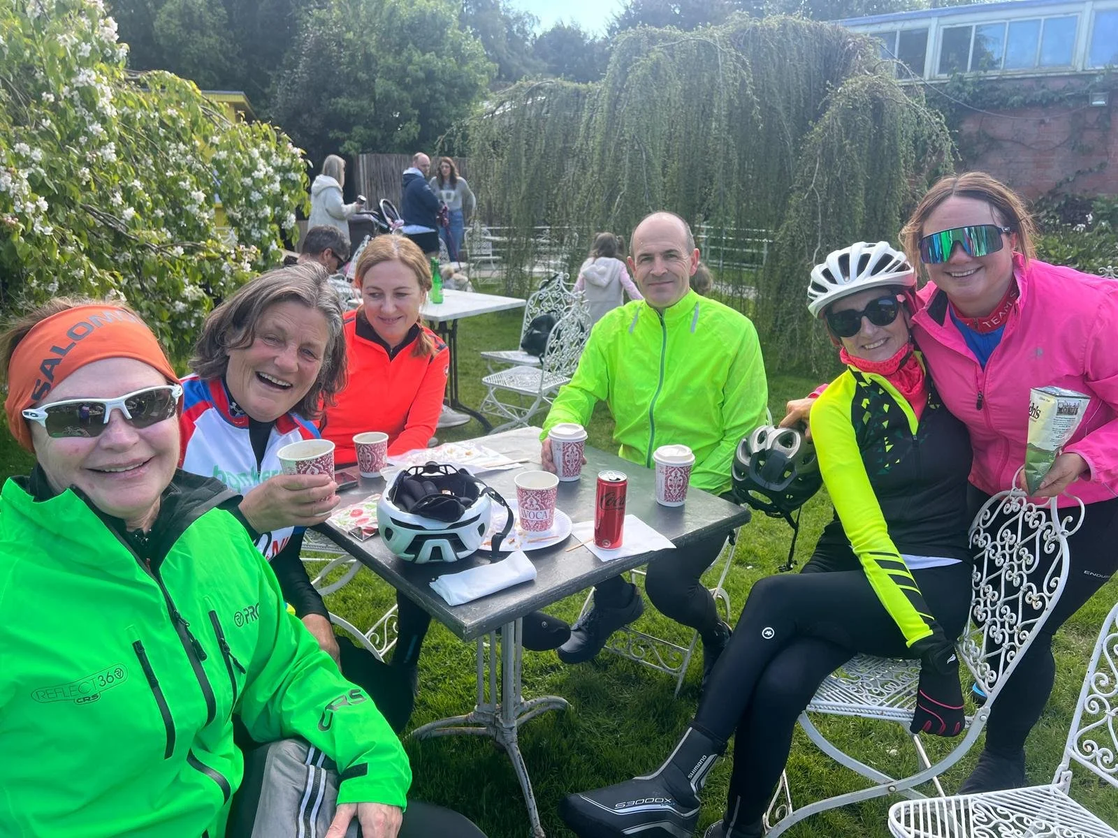 Group of six cyclists sitting and standing at an outdoor table with green foliage and other people in the background. They are smiling, with some wearing cycling helmets and sunglasses, holding cups and snacks.