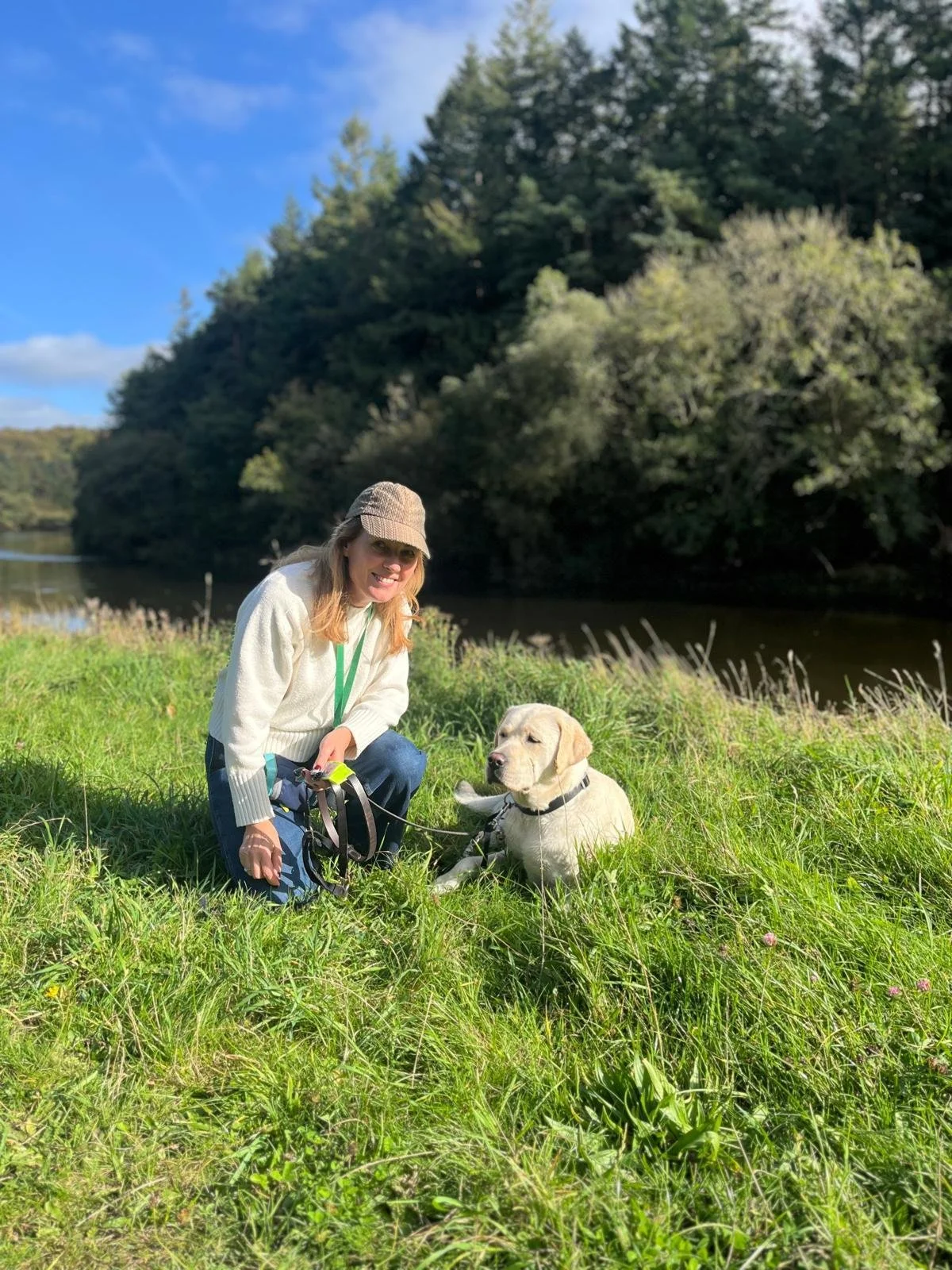 A woman with a yellow Labrador retriever puppy sitting on green grass near a river, with trees and a blue sky in the background.