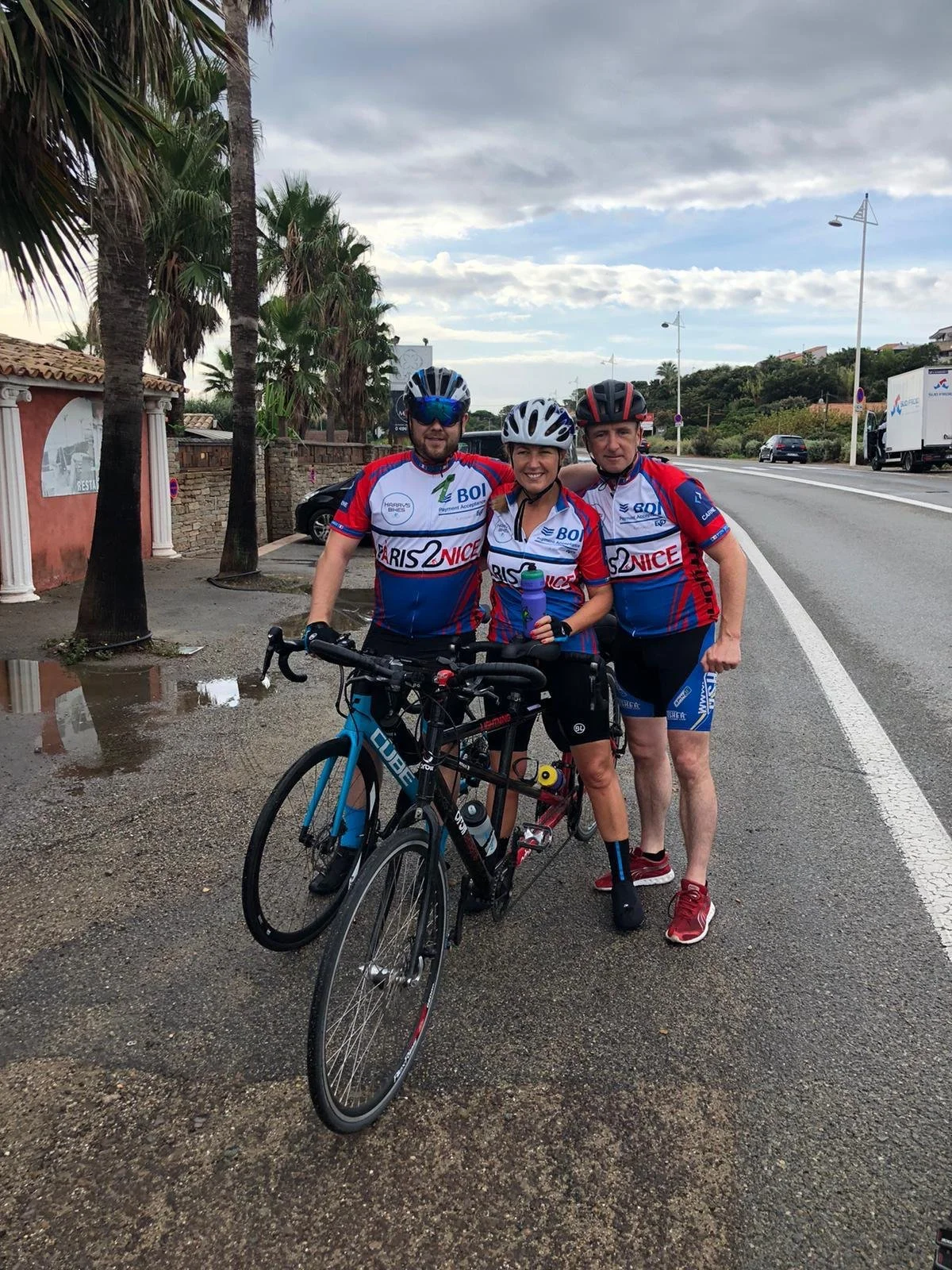 Three cyclists in matching blue and red jerseys standing with bikes on a wet road near palm trees, with a cloudy sky overhead.