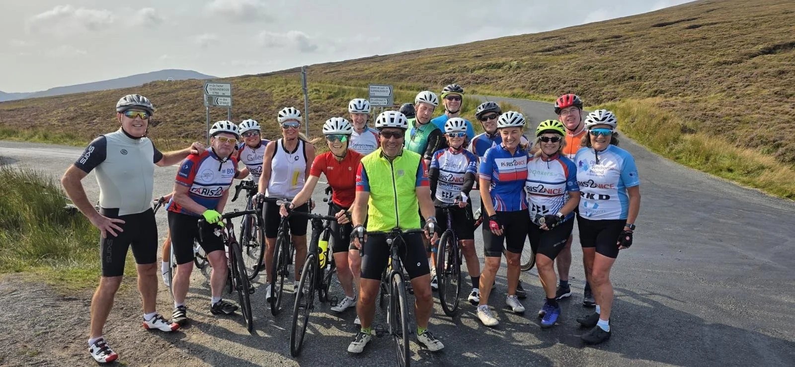 A group of cyclists wearing helmets and cycling gear standing on a rural road, with rolling hills in the background.