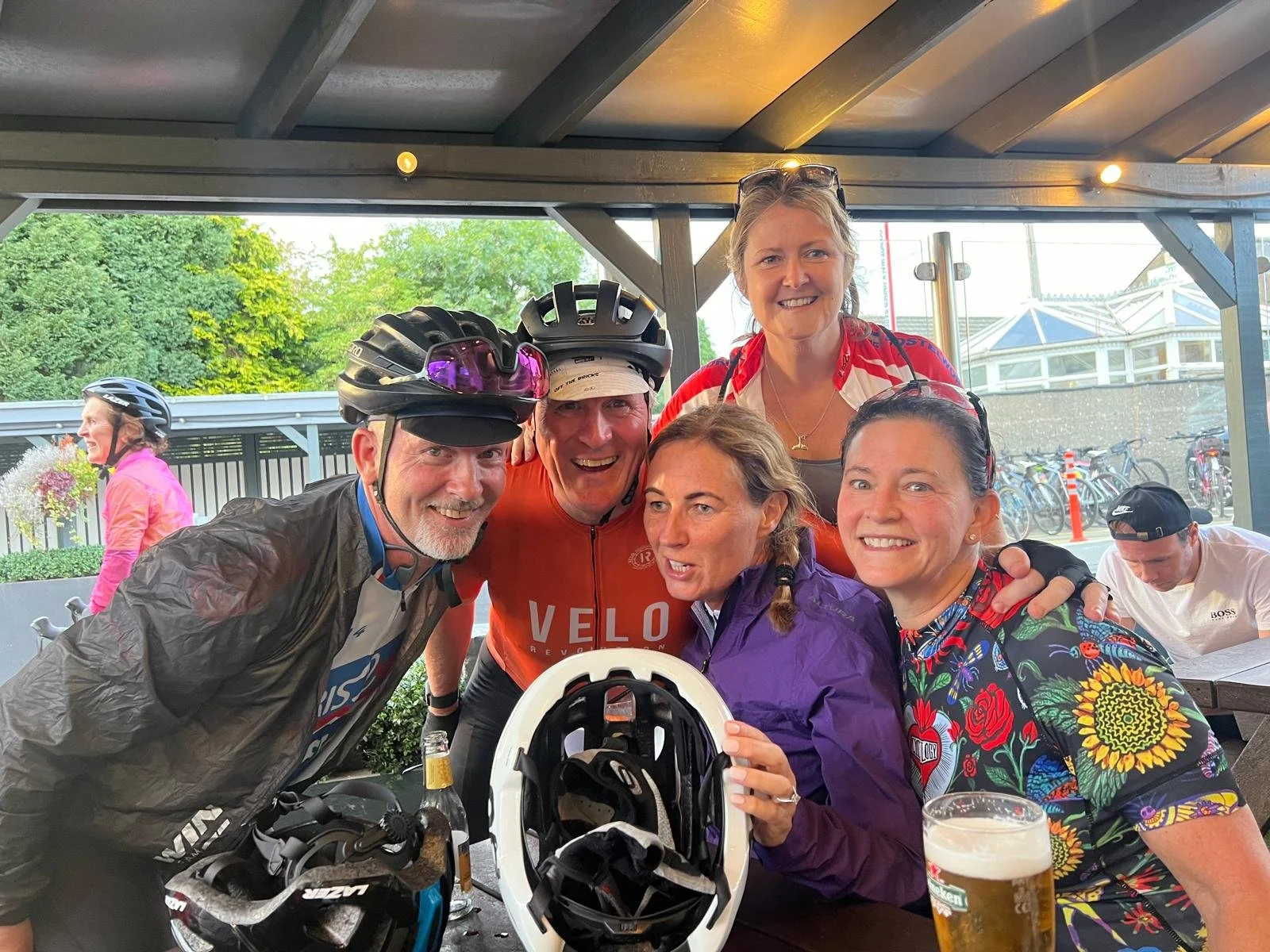 A group of six people, with some wearing cycling helmets, gathered around a table at an indoor or covered outdoor space, smiling and celebrating. One person is holding a bicycle helmet, at least two people are wearing sunglasses, and there is a glass