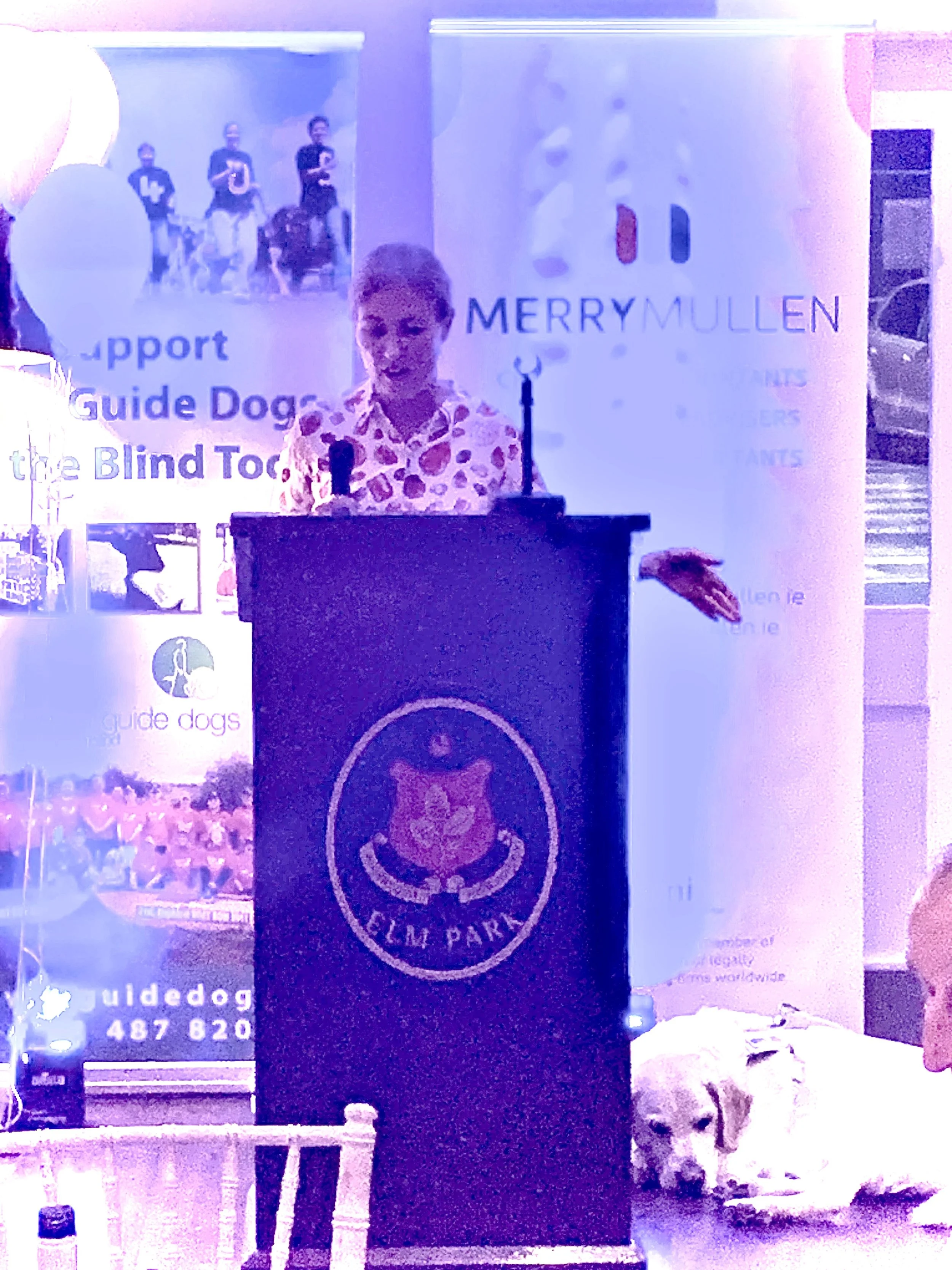 Woman speaking at a podium with a guide dog lying beside her, event banners in the background for Guide Dogs and MERRY MULLEN.