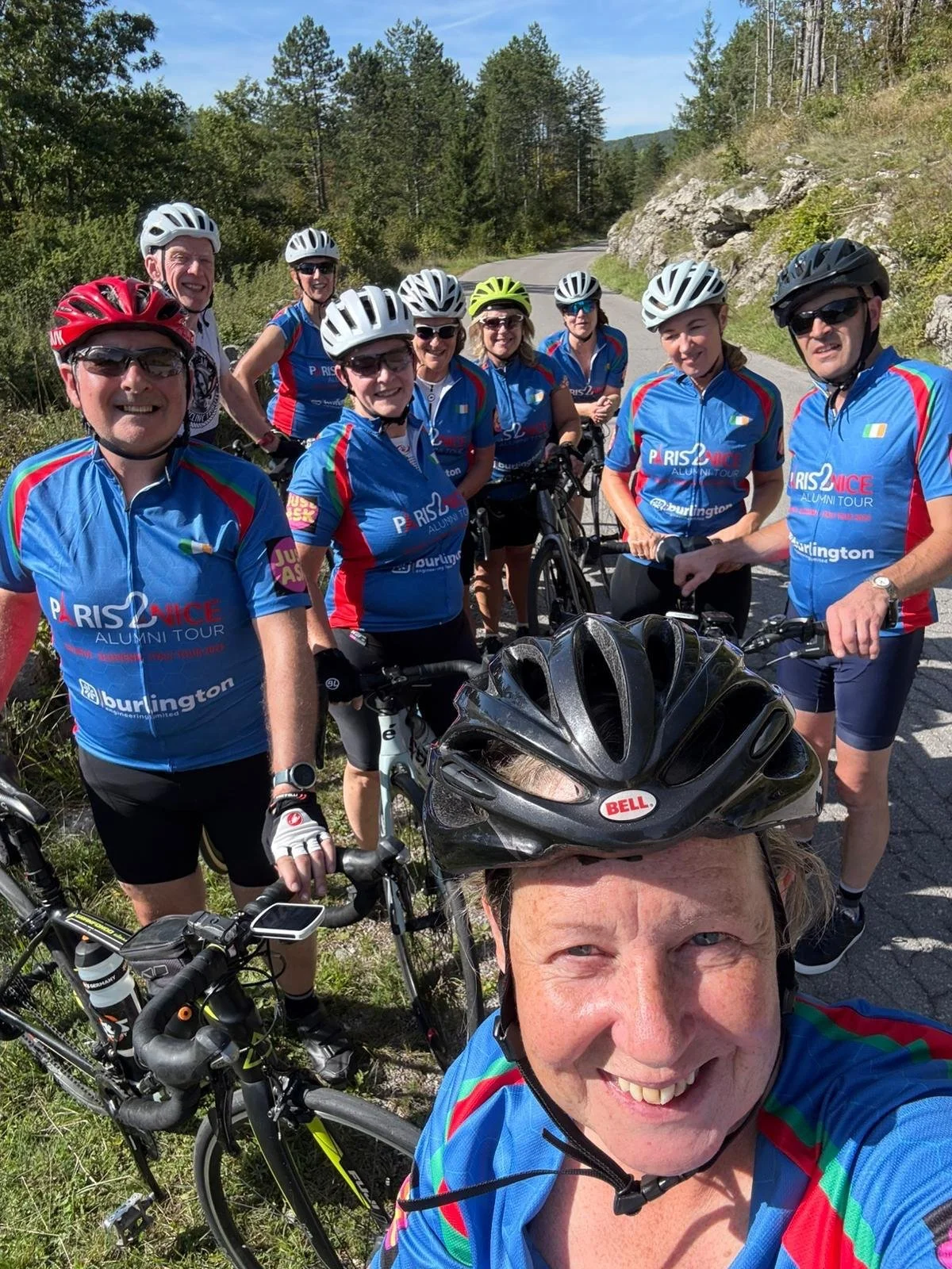 Group of nine cyclists in blue jerseys taking a selfie on a mountain road surrounded by trees and rocks.