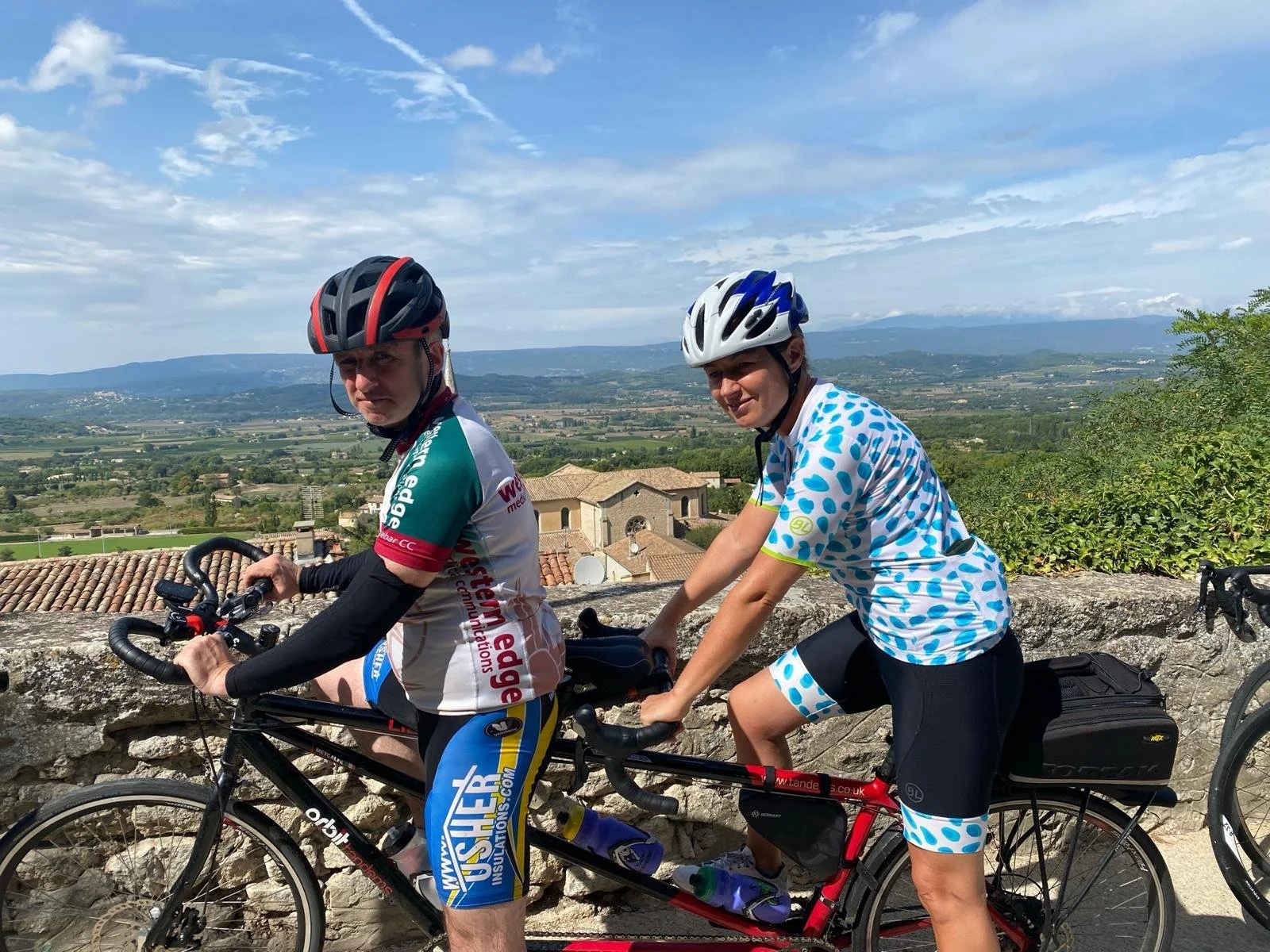 Two cyclists wearing helmets and sports clothing, riding on a road with a scenic countryside view in the background.