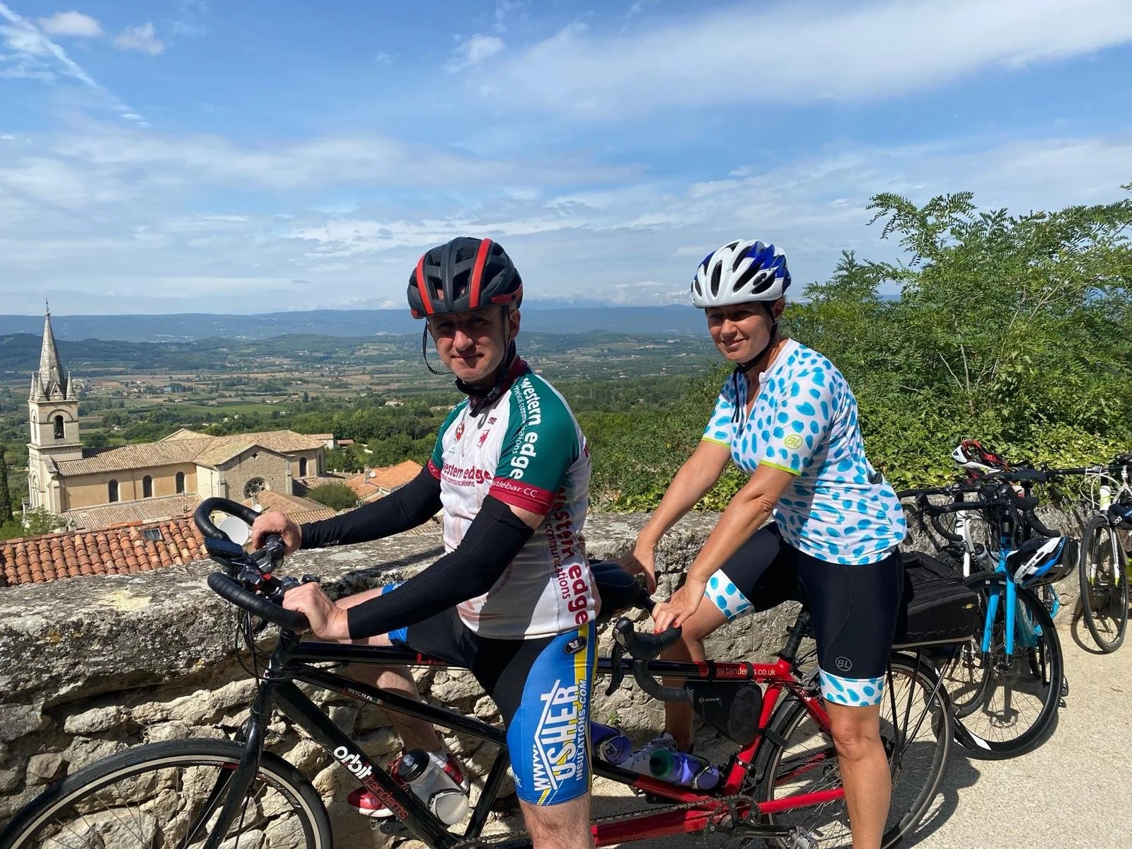 Two cyclists wearing helmets and jerseys on a tandem bike, with rolling hills, a small church, and blue sky in the background.
