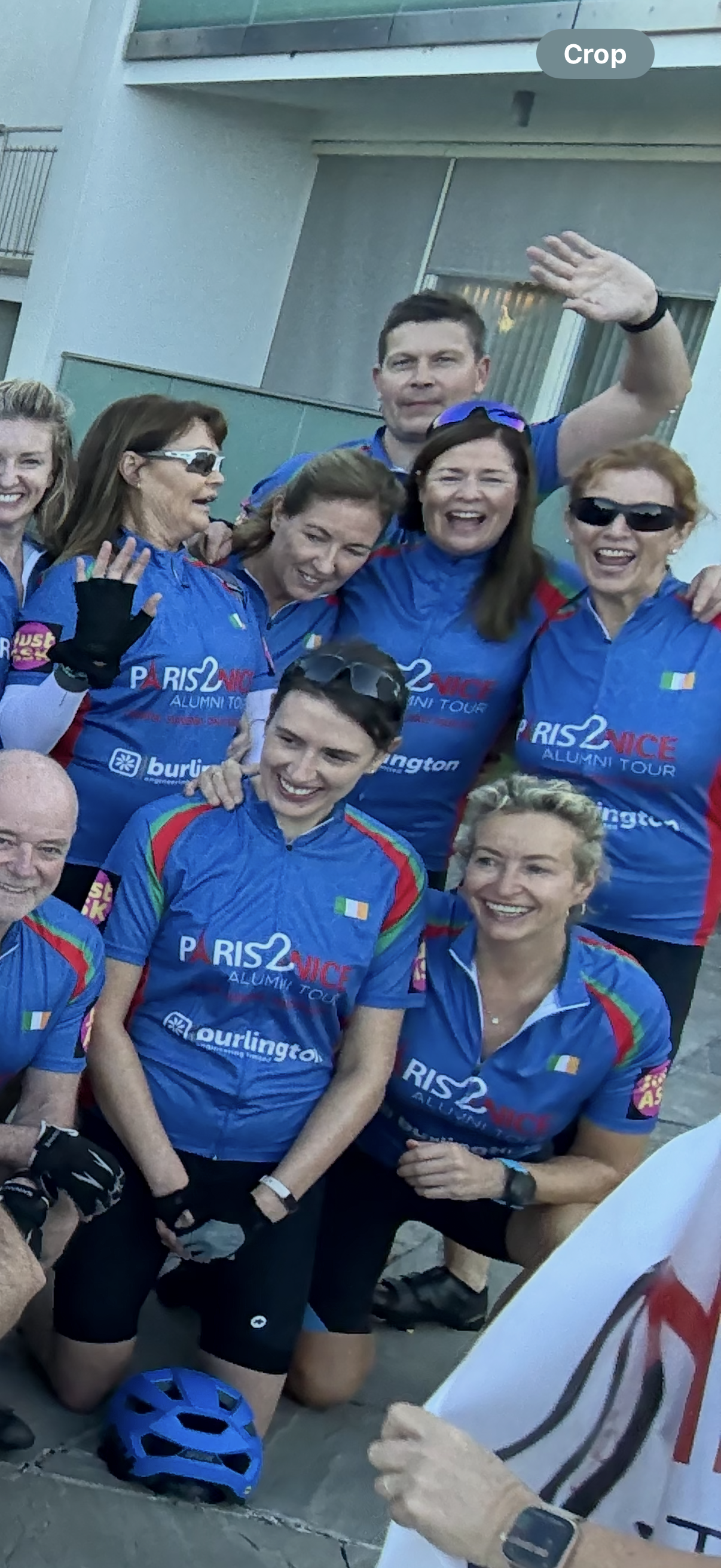 Group of people in blue cycling jerseys posing for a photo. Some are smiling and waving, and they appear to be celebrating after a cycling event.
