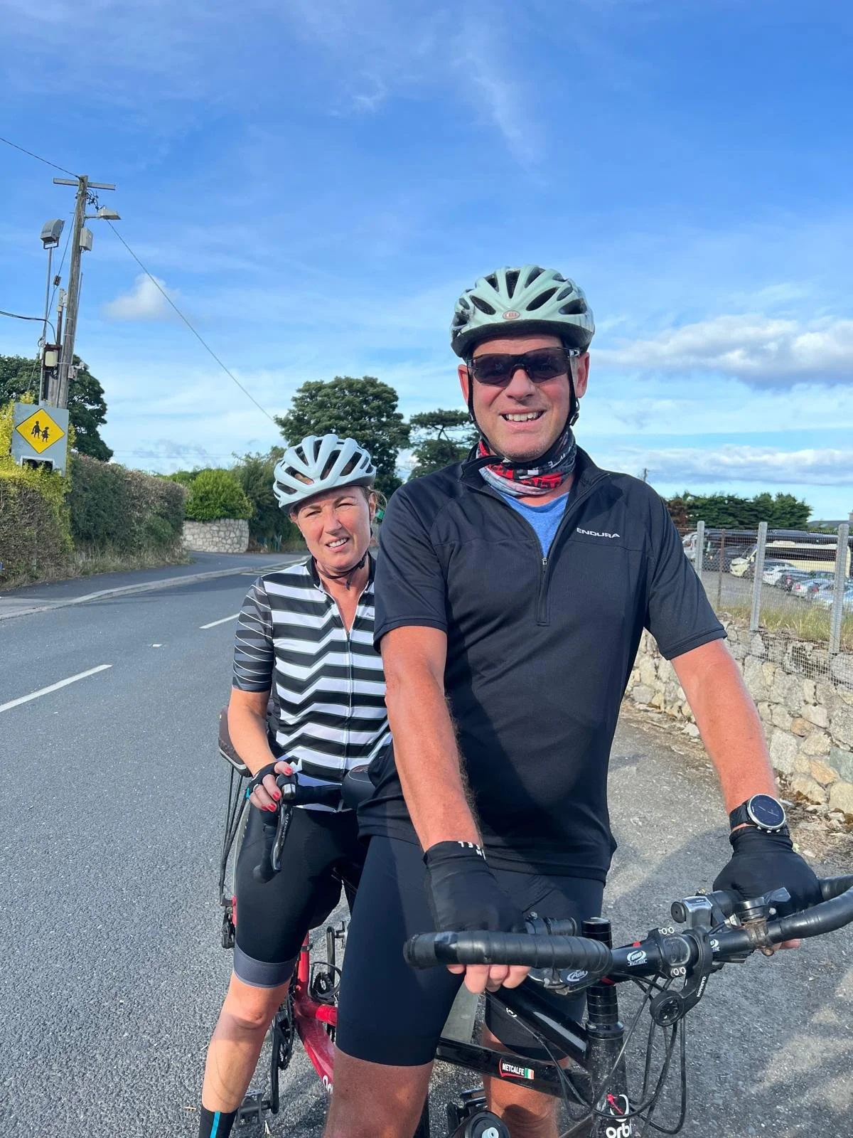 A man and woman wearing cycling helmets and gear, riding bikes on a paved road on a sunny day.