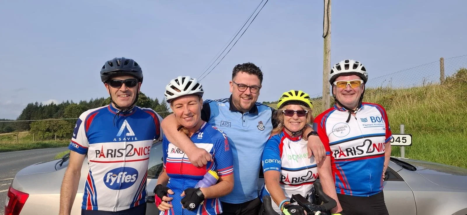 Five cyclists wearing colorful jerseys and helmets, standing together outdoors next to a silver car, smiling at the camera with blue sky and grassy hillside in the background.