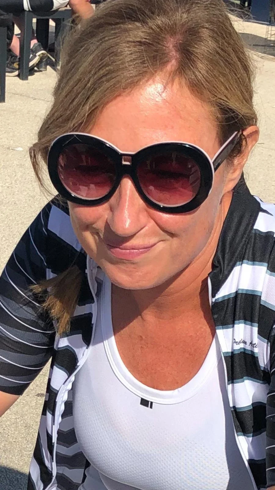 A woman wearing large black sunglasses, a black and white striped cycling jersey, and a white athletic shirt is smiling outdoors on a sunny day.