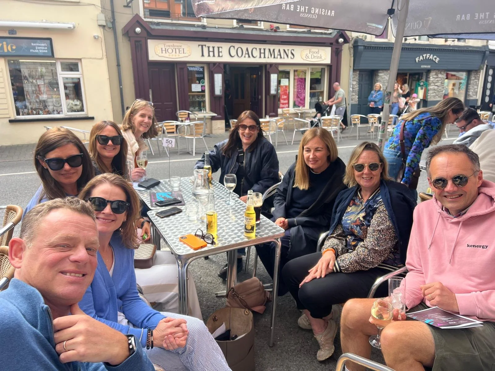 Group of ten friends sitting at an outdoor cafe table on the street, smiling, with some holding drinks, during daytime.