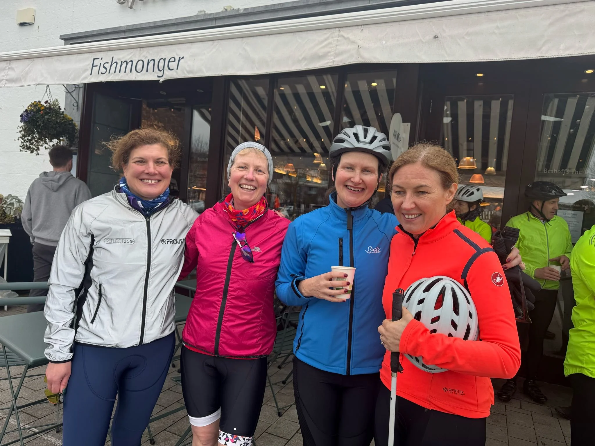 Four women in cycling gear smiling outside a cafe, some holding helmets and drinks, with several people in bright jackets in the background.