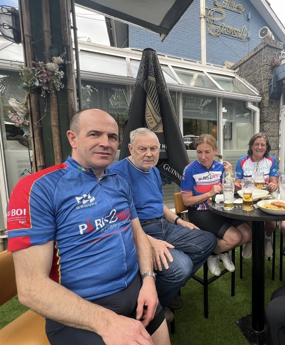 Four people sitting at an outdoor table, two women and two men, wearing cycling jerseys, with drinks and food on the table, outside a building with a sign that reads "The Goat."