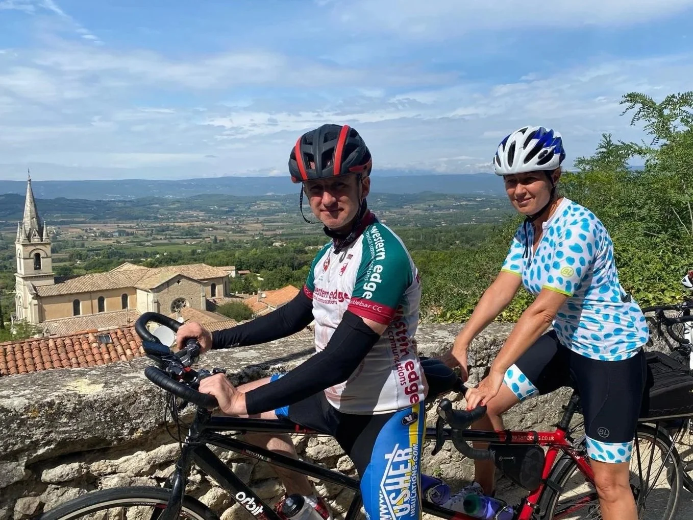 Two cyclists, a man and a woman, wearing helmets and cycling jerseys, pose with their bikes on a hilltop overlooking a town with a church and a landscape of fields and hills in the background on a partly cloudy day.