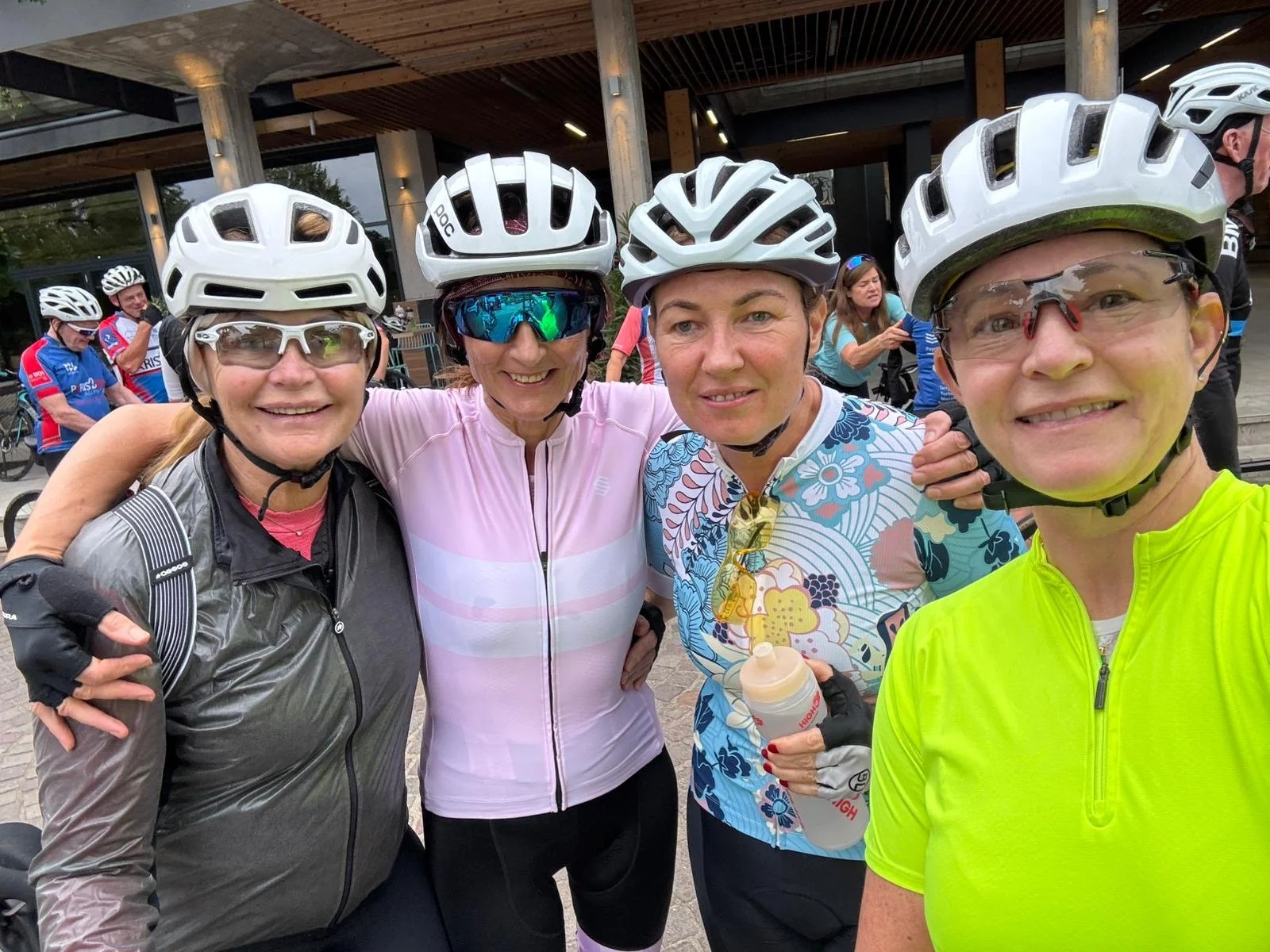 Four women in cycling gear and helmets posing for a photo, smiling, with other cyclists in the background.