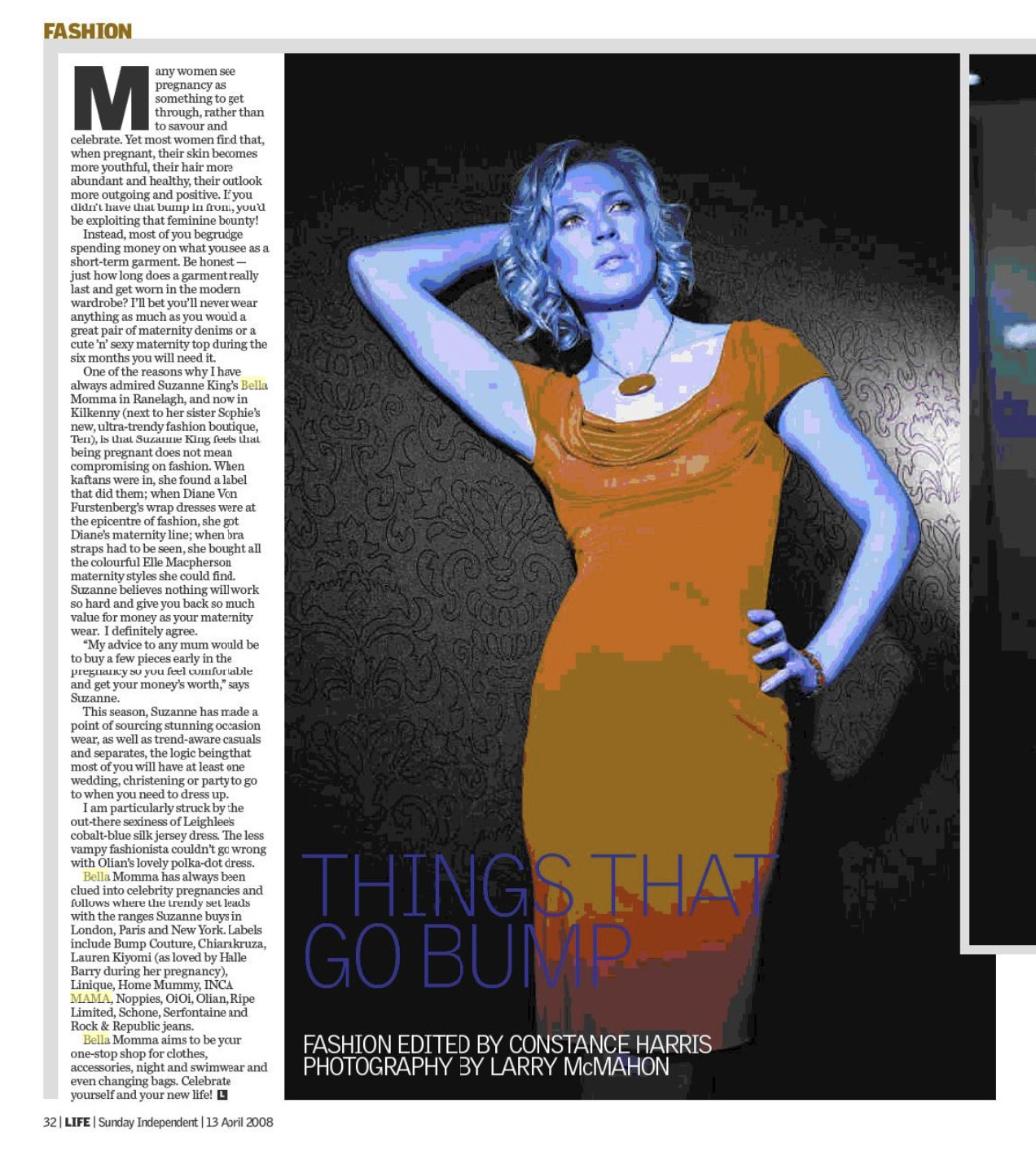 A woman with blonde curly hair in an orange dress standing against a dark patterned background with her hand on her hip, posing confidently.