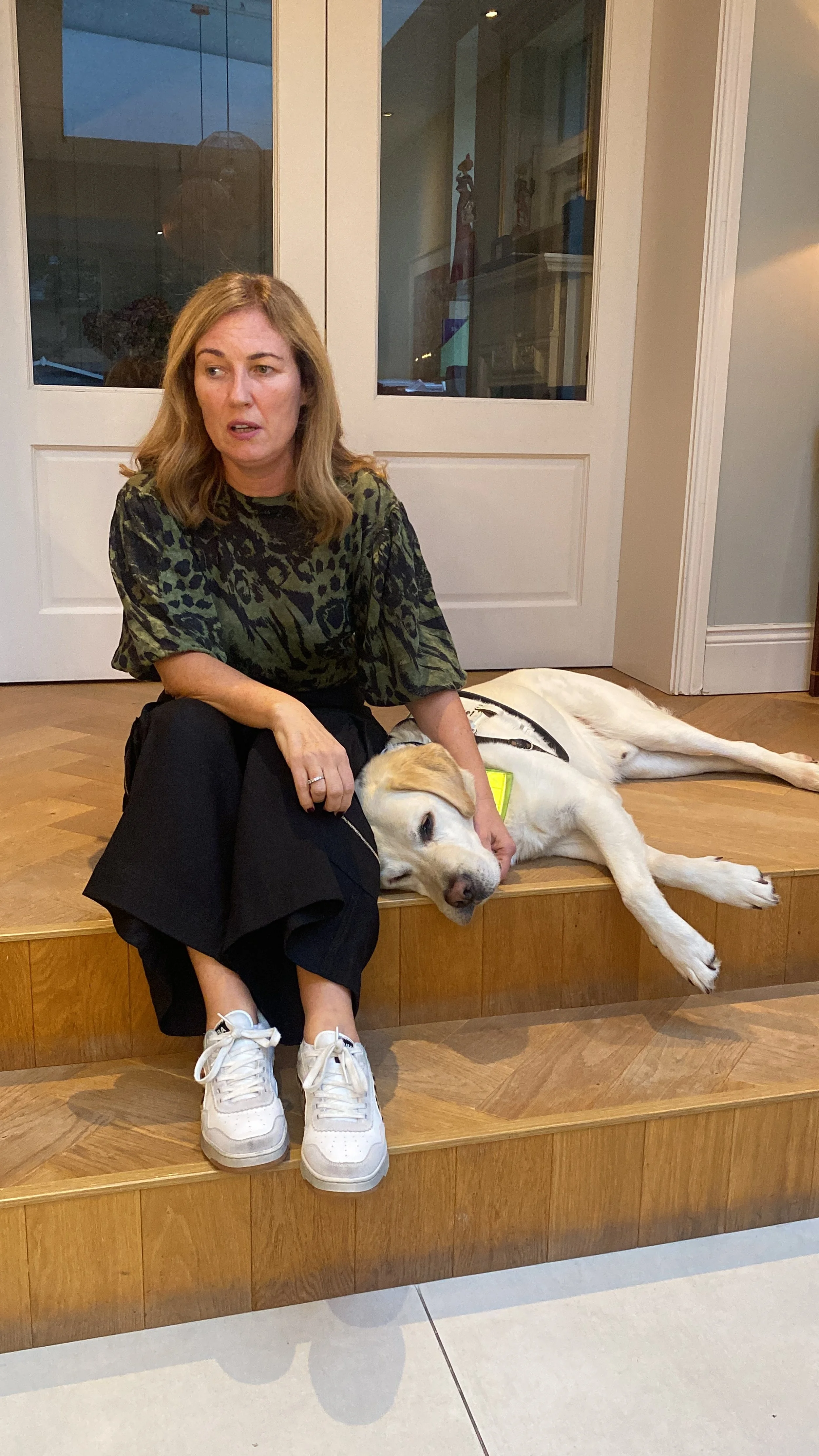 A woman sitting on wooden steps with a tired-looking Labrador Retriever lying next to her. The woman has shoulder-length blonde hair and is wearing a green and black patterned top and white sneakers. The dog is wearing a harness and is lying on its side with its head resting on the woman's lap, in front of a white door with glass panes.