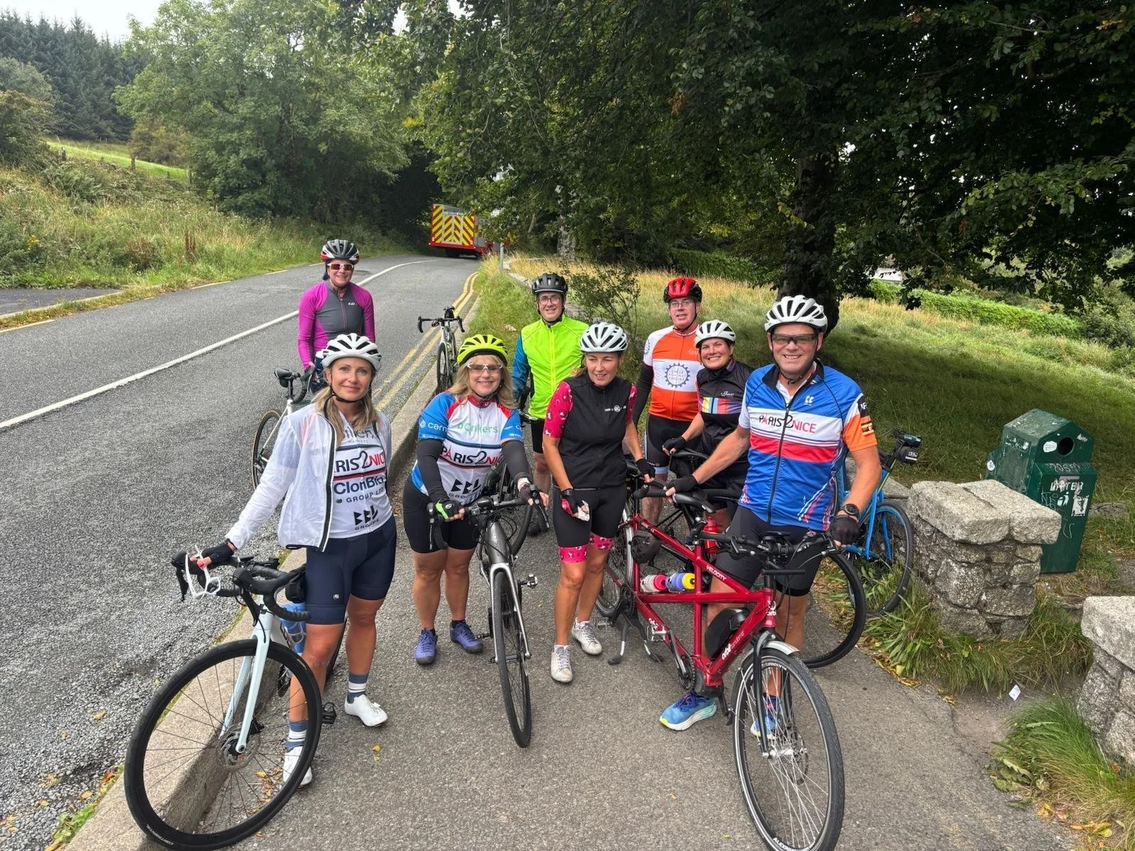 Group of nine cyclists with helmets and cycling gear, standing with their bikes on a rural road with greenery and trees, and a fire truck in the background.