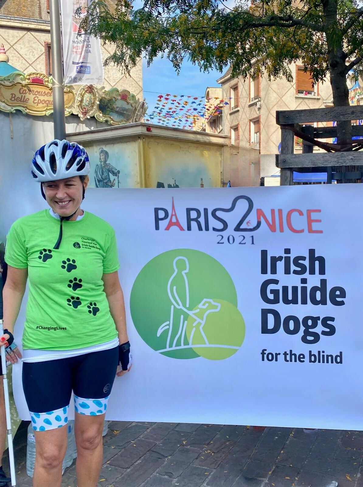 A woman in cycling gear and a helmet standing next to a large sign that reads 'Paris2Nice 2021 Irish Guide Dogs for the Blind' with a logo of a person walking a guide dog, outdoors in a lively, decorated area.