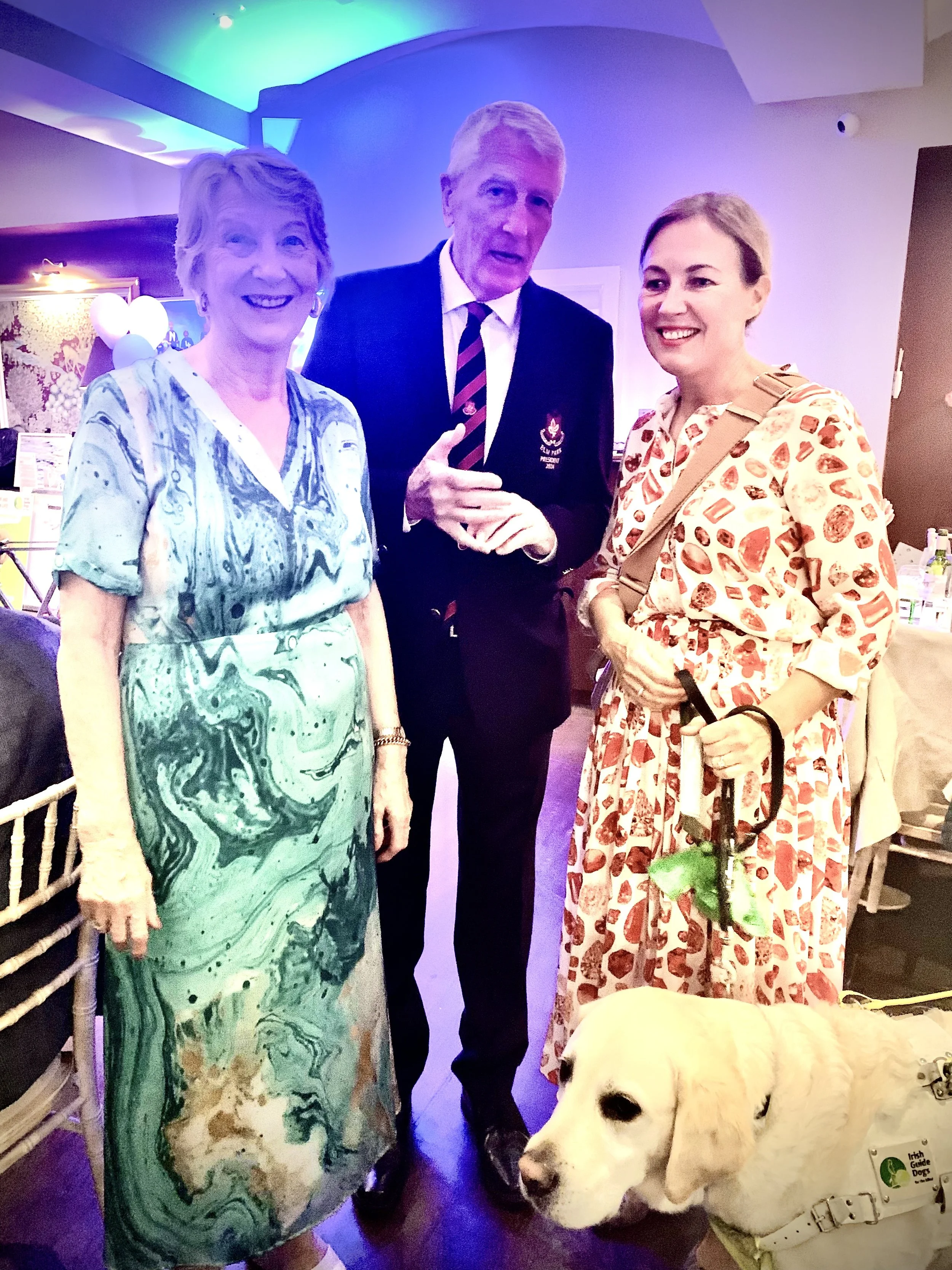 Three women and a man standing together indoors with a Labrador Guide Dog. The women are smiling, and the man is making a hand gesture. The setting appears to be a social event with colorful lighting and tables in the background.