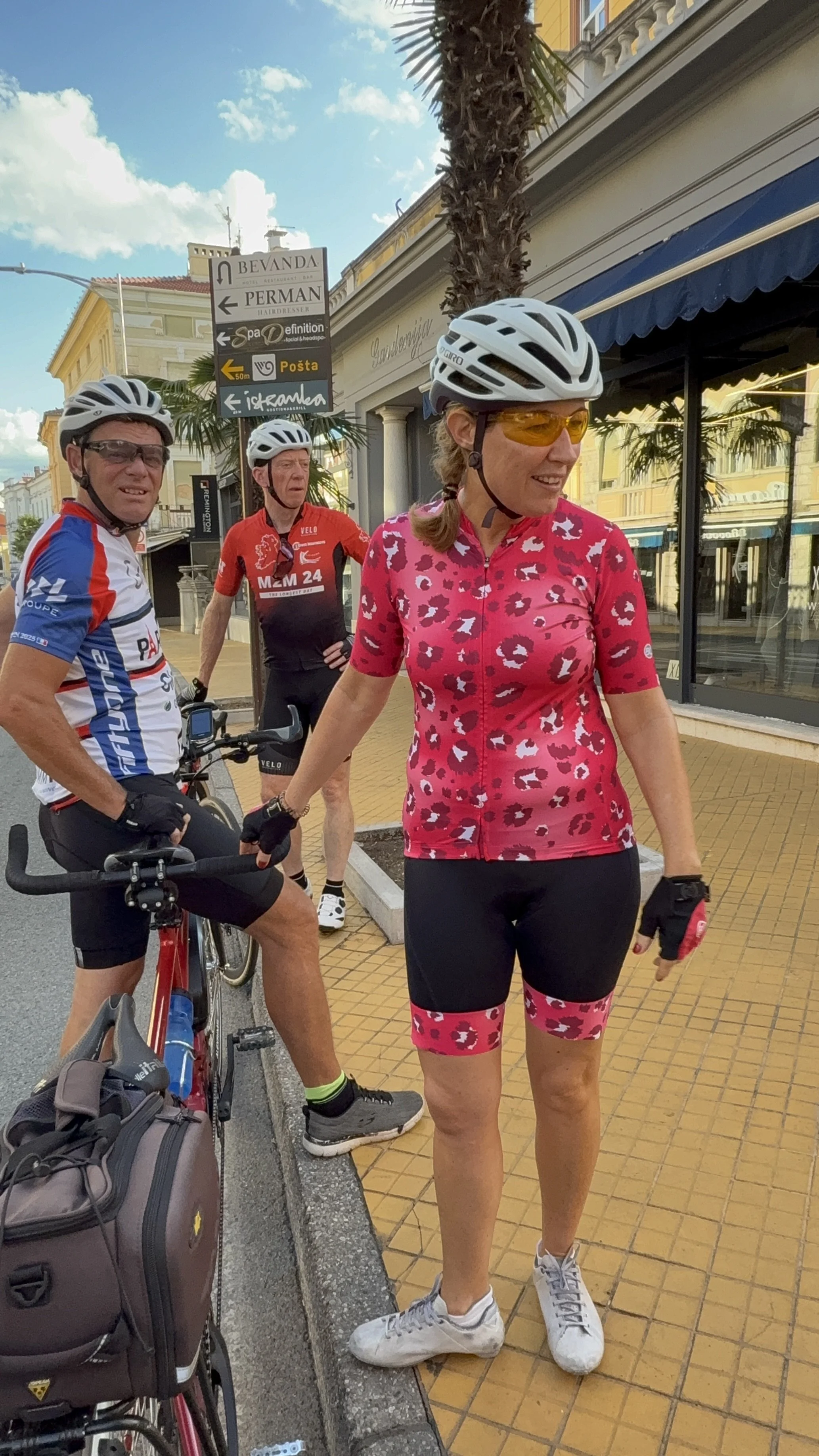 Three cyclists in colorful gear stand on a city sidewalk. One woman in a pink leopard-print cycling outfit is smiling, wearing yellow tinted glasses and a helmet. Two men, one in a blue and white jersey and the other in a red and black kit, stand nea