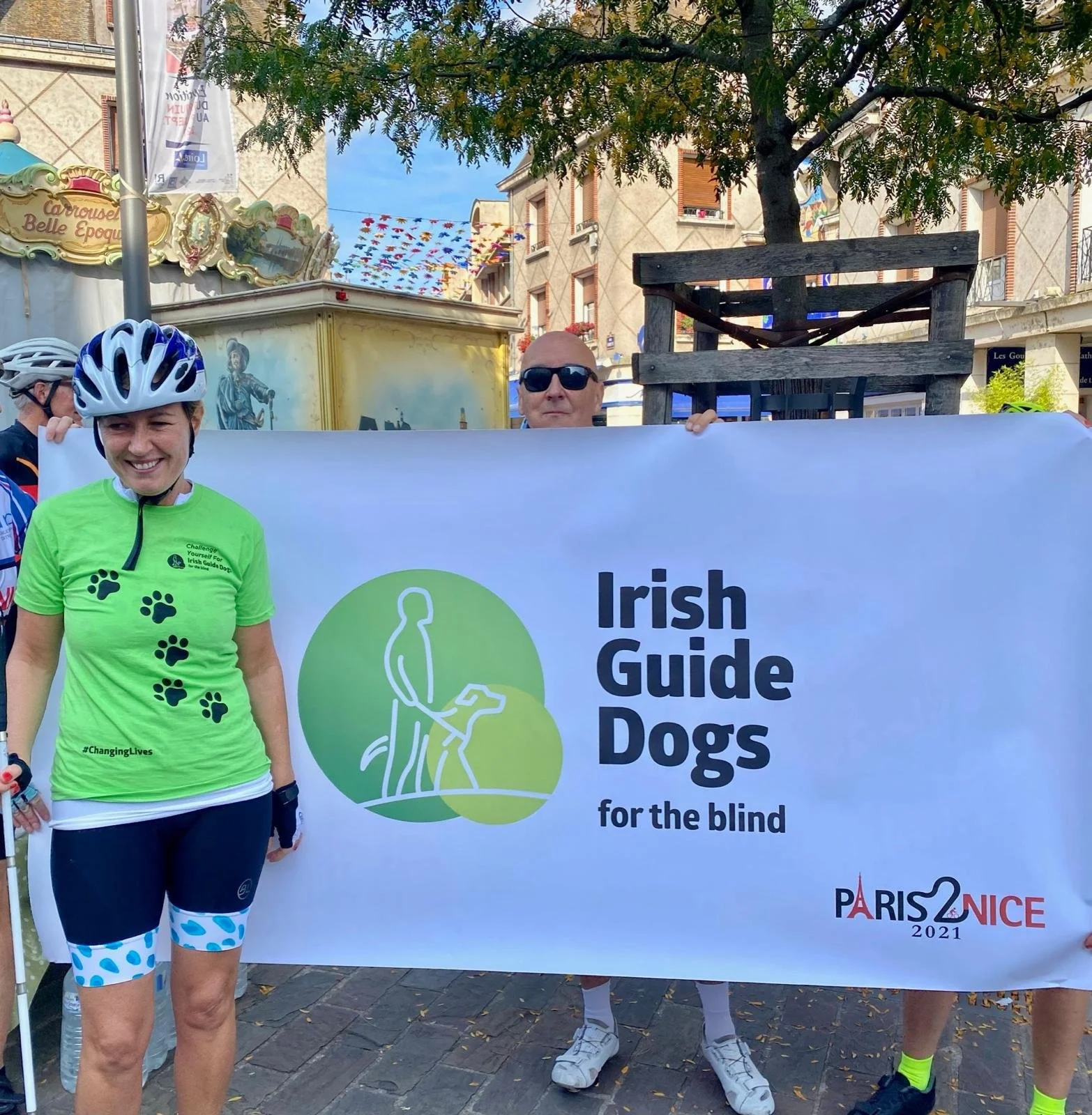 People holding a banner that reads 'Irish Guide Dogs for the blind' during an outdoor event. One woman wearing a green shirt with paw prints and cycling gear, including a helmet, stands to the left. Two individuals hold the banner, with one in sungla